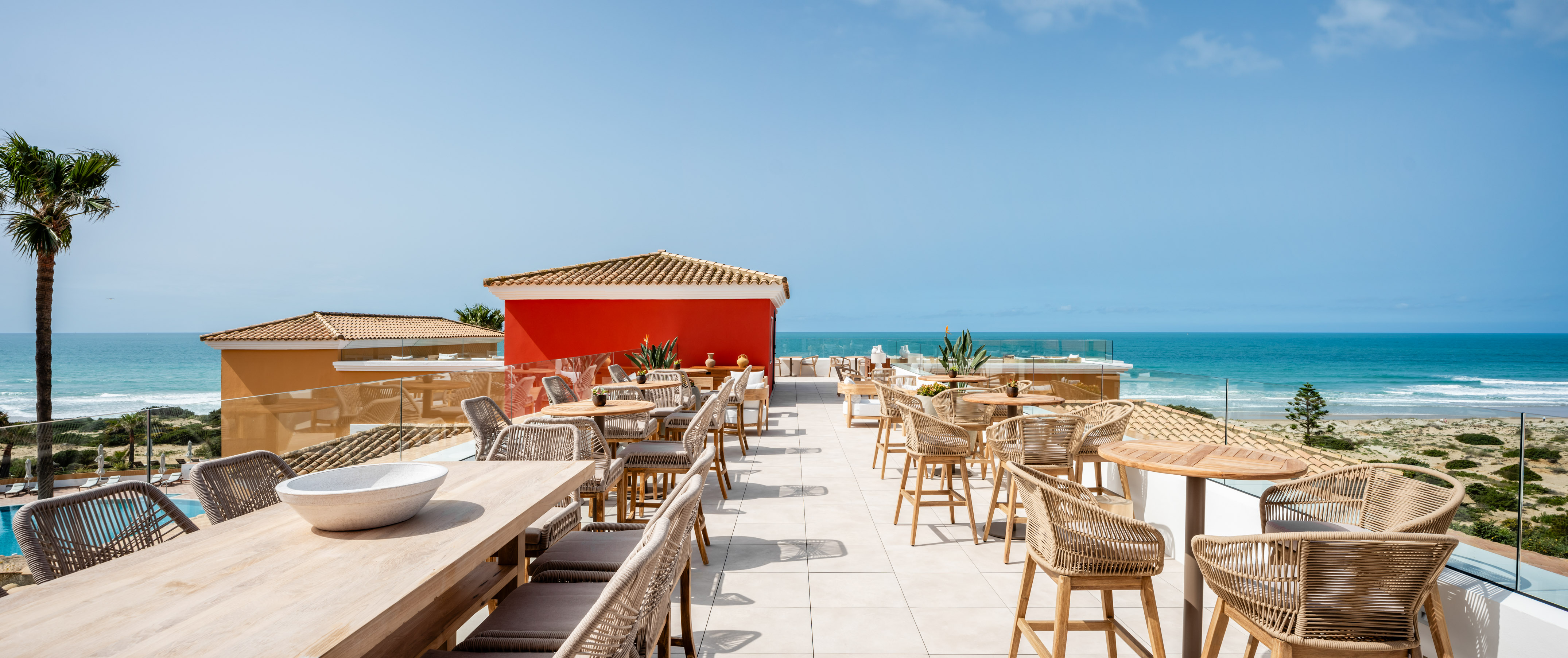 Oceanfront terrace restaurant with woven furniture, blue sea, and red building.