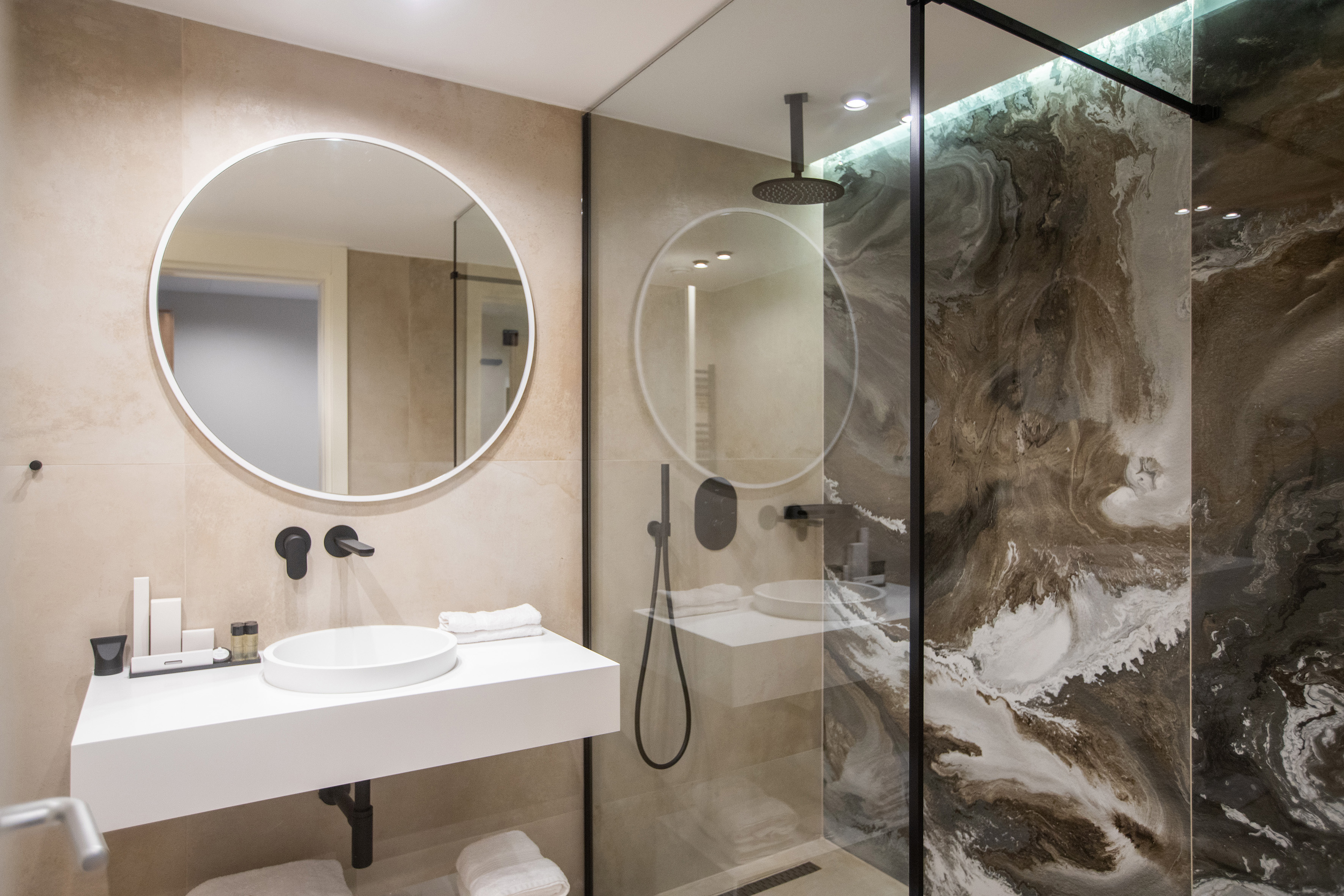 Minimalist bathroom with round mirror, white floating vanity, and marble-effect shower.
