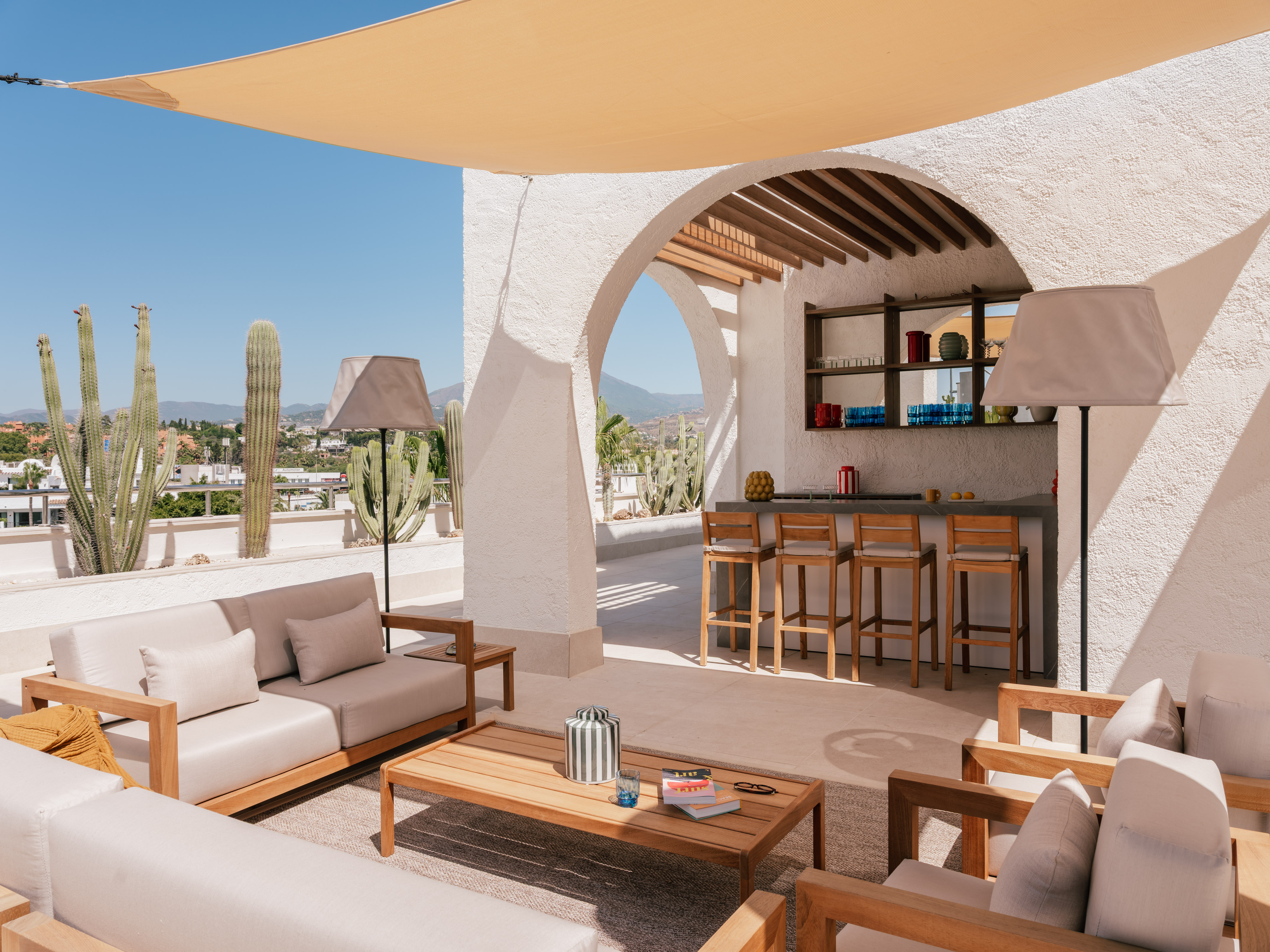 Sunny rooftop terrace with outdoor lounge, bar, cacti, and white stucco arches.