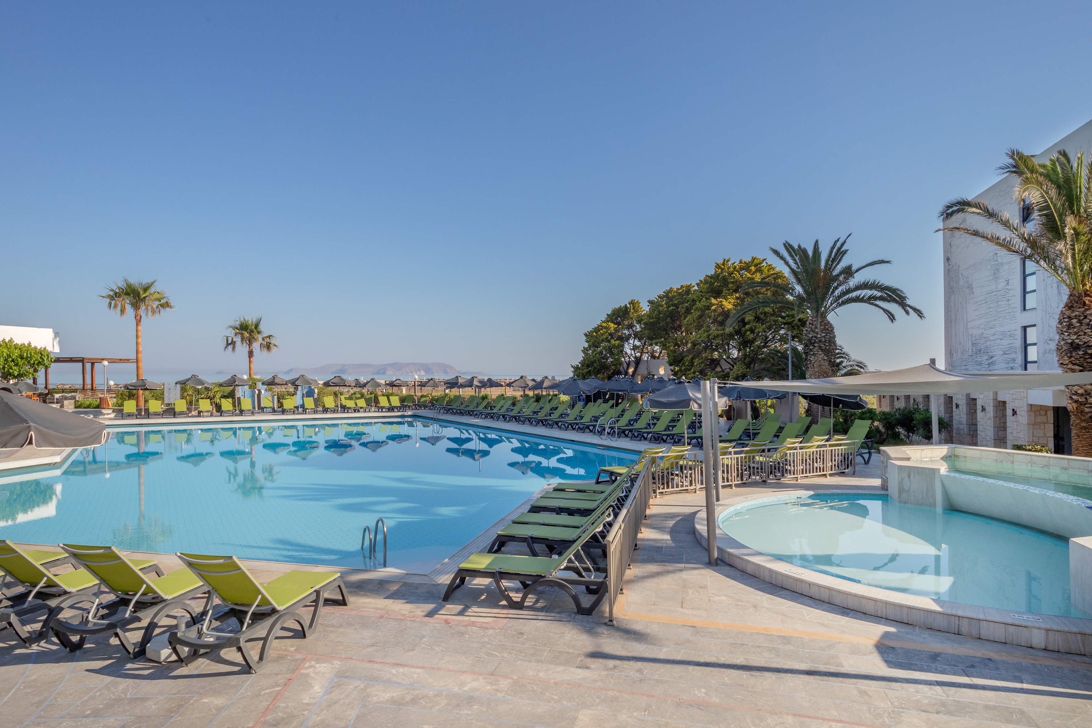 Sunny resort pool with green loungers, palm trees, and distant sea view.