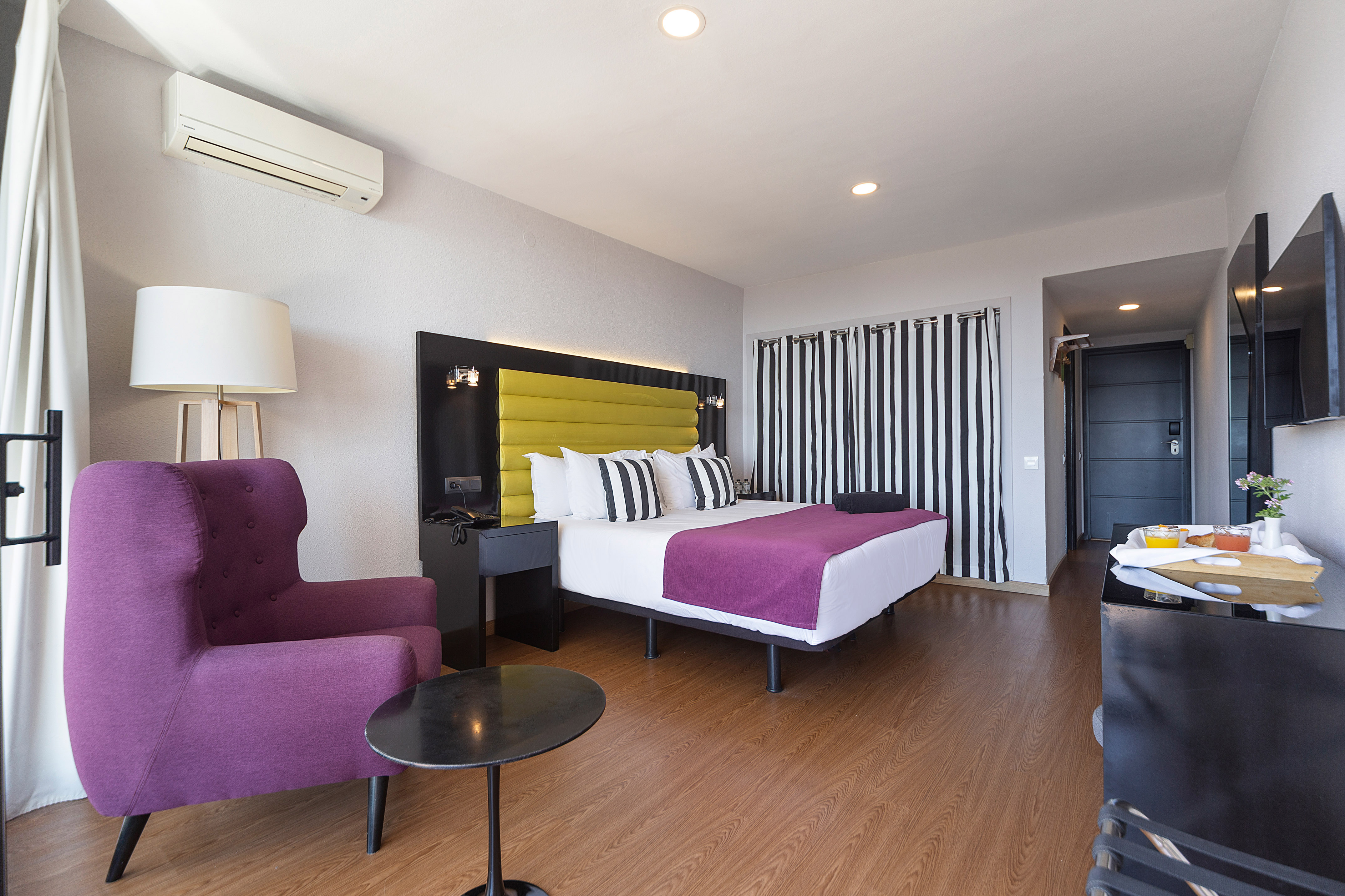 Modern hotel bedroom with yellow headboard, purple armchair, and breakfast tray.