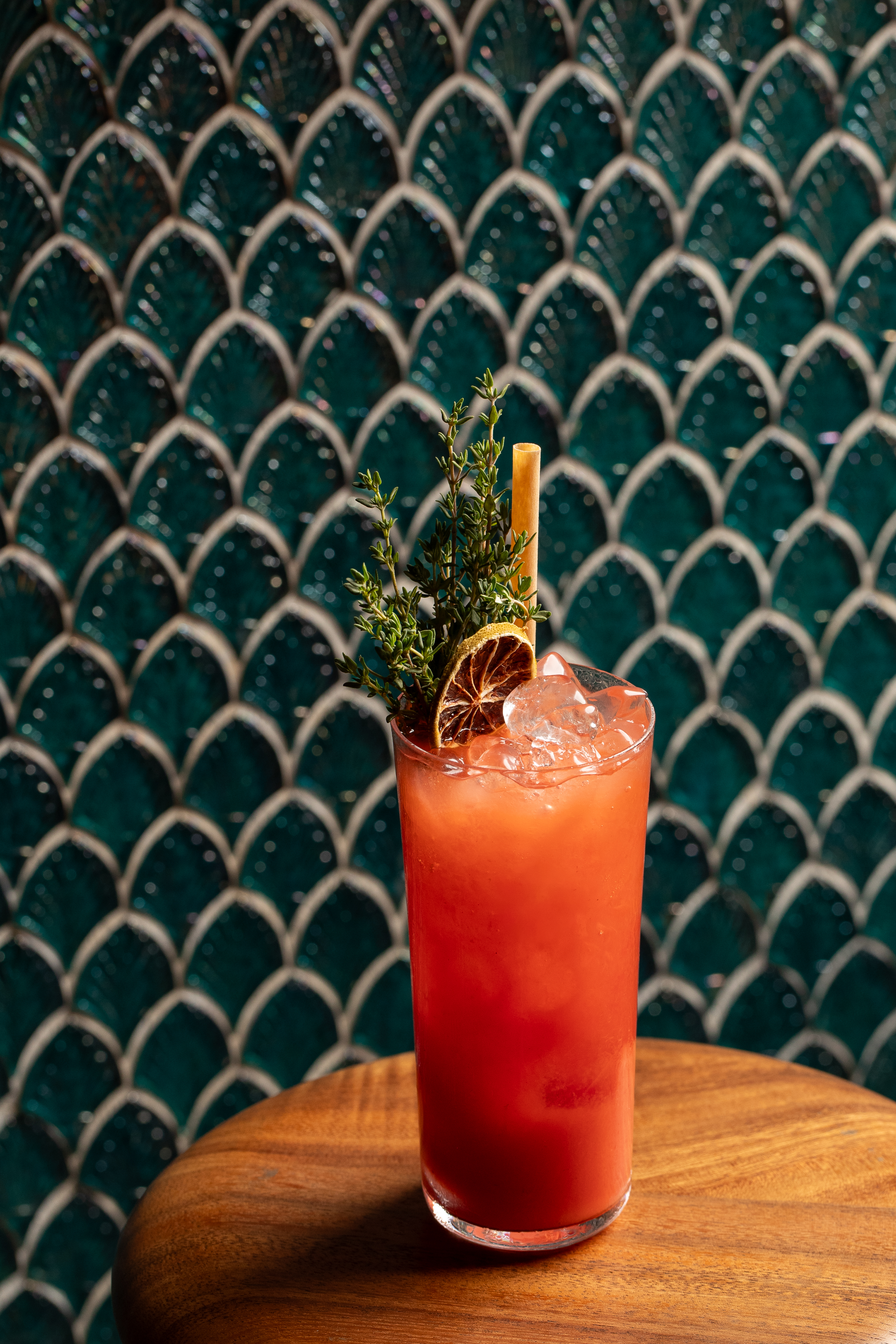 Red cocktail with dried orange and thyme garnish, against green scale tiles.