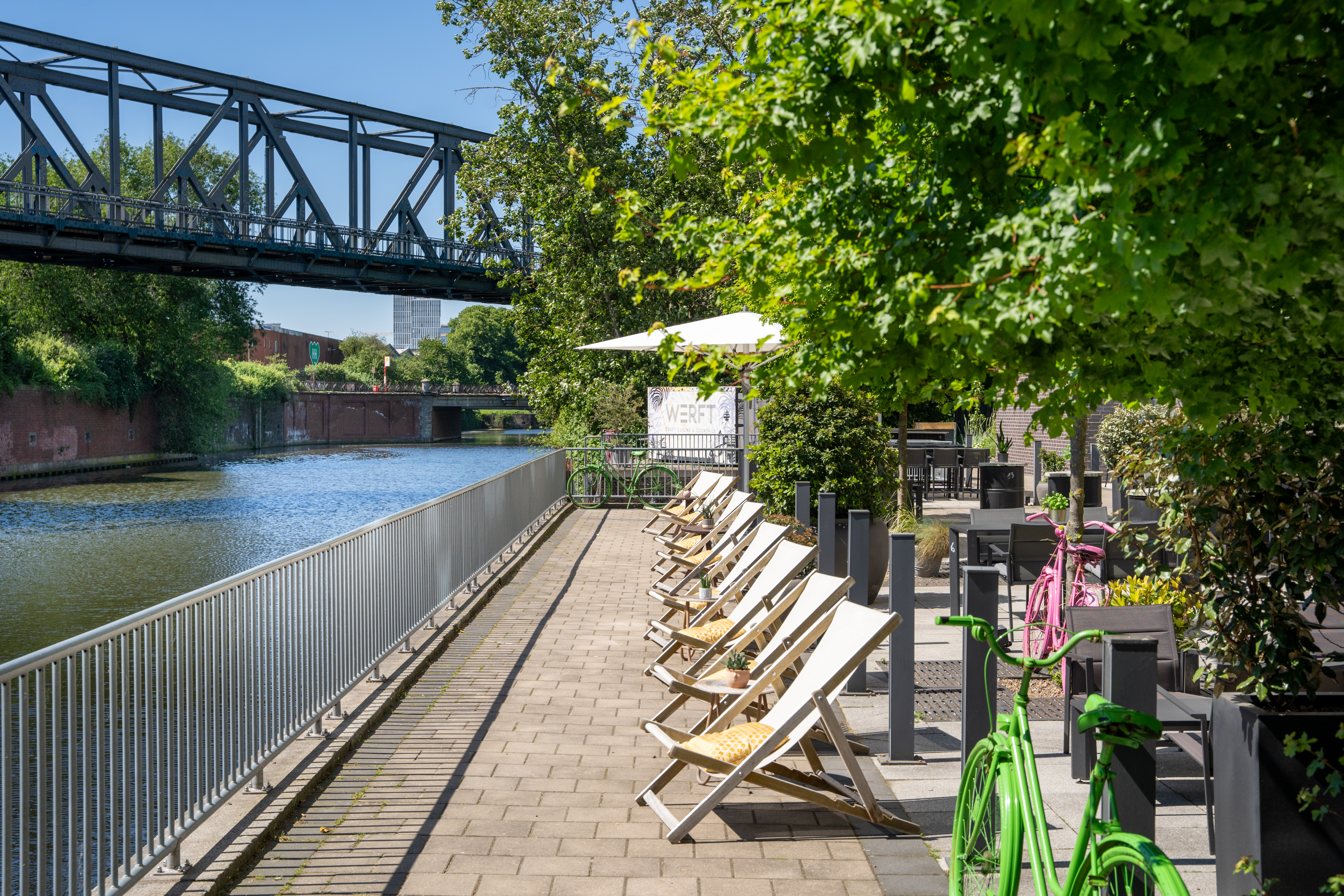 Waterfront cafe patio with deck chairs, river, green trees, and metal bridge.