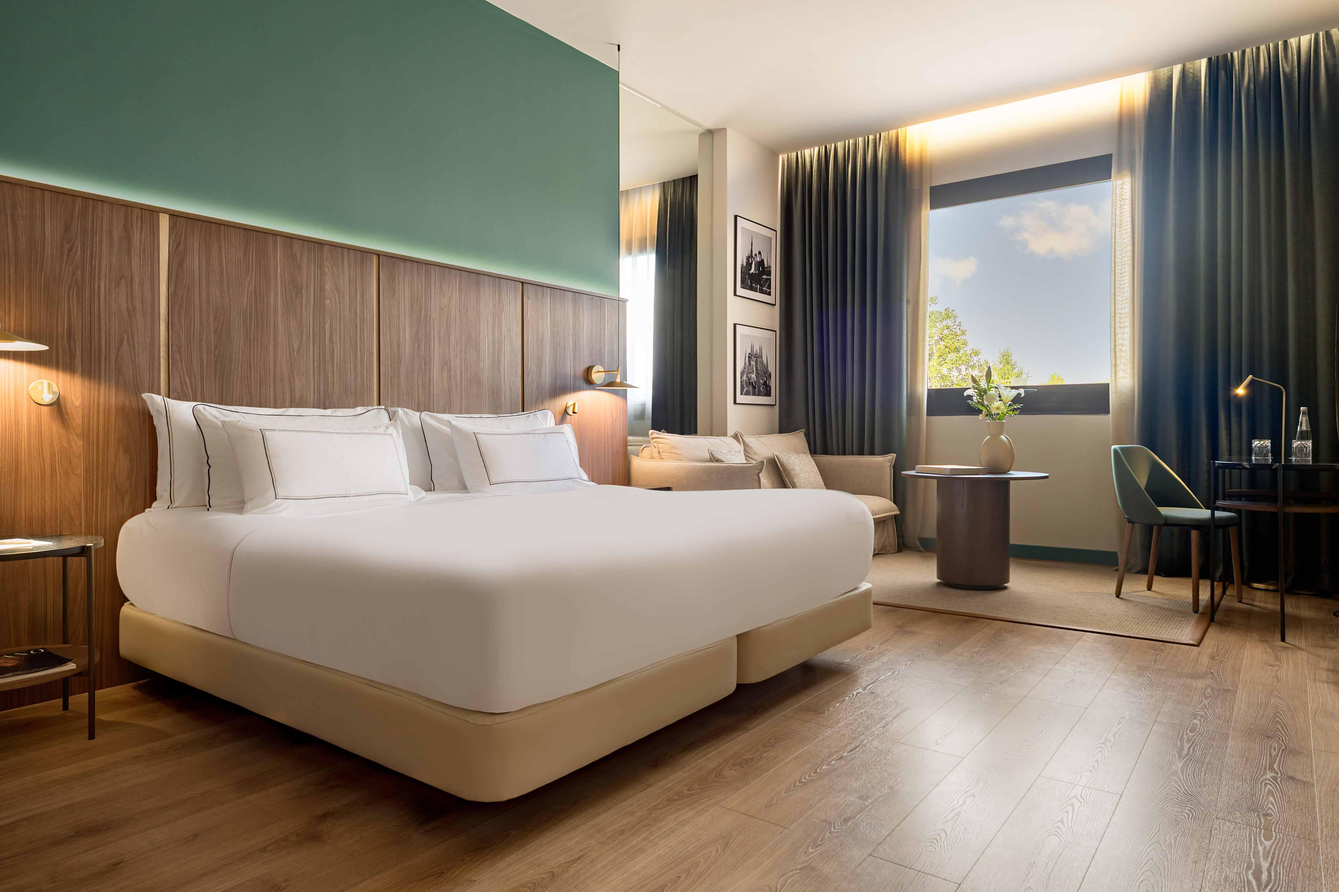 Modern guest room with king bed, green accent wall, wood floor, and large window.