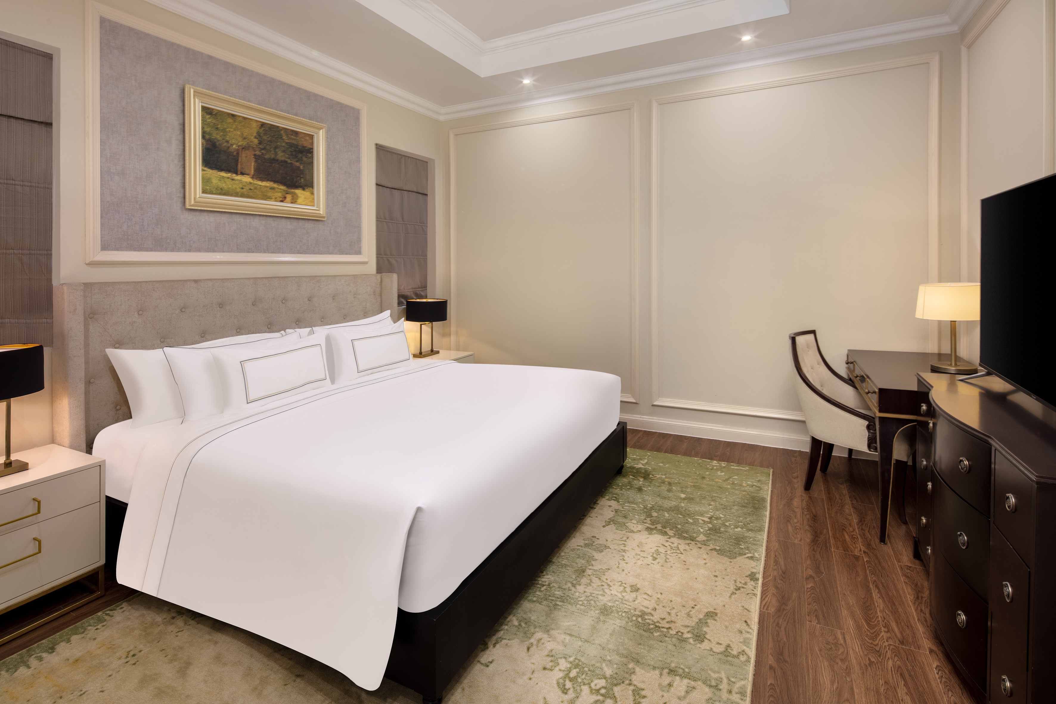 Hotel bedroom with king bed, tufted headboard, desk, TV, and patterned rug.
