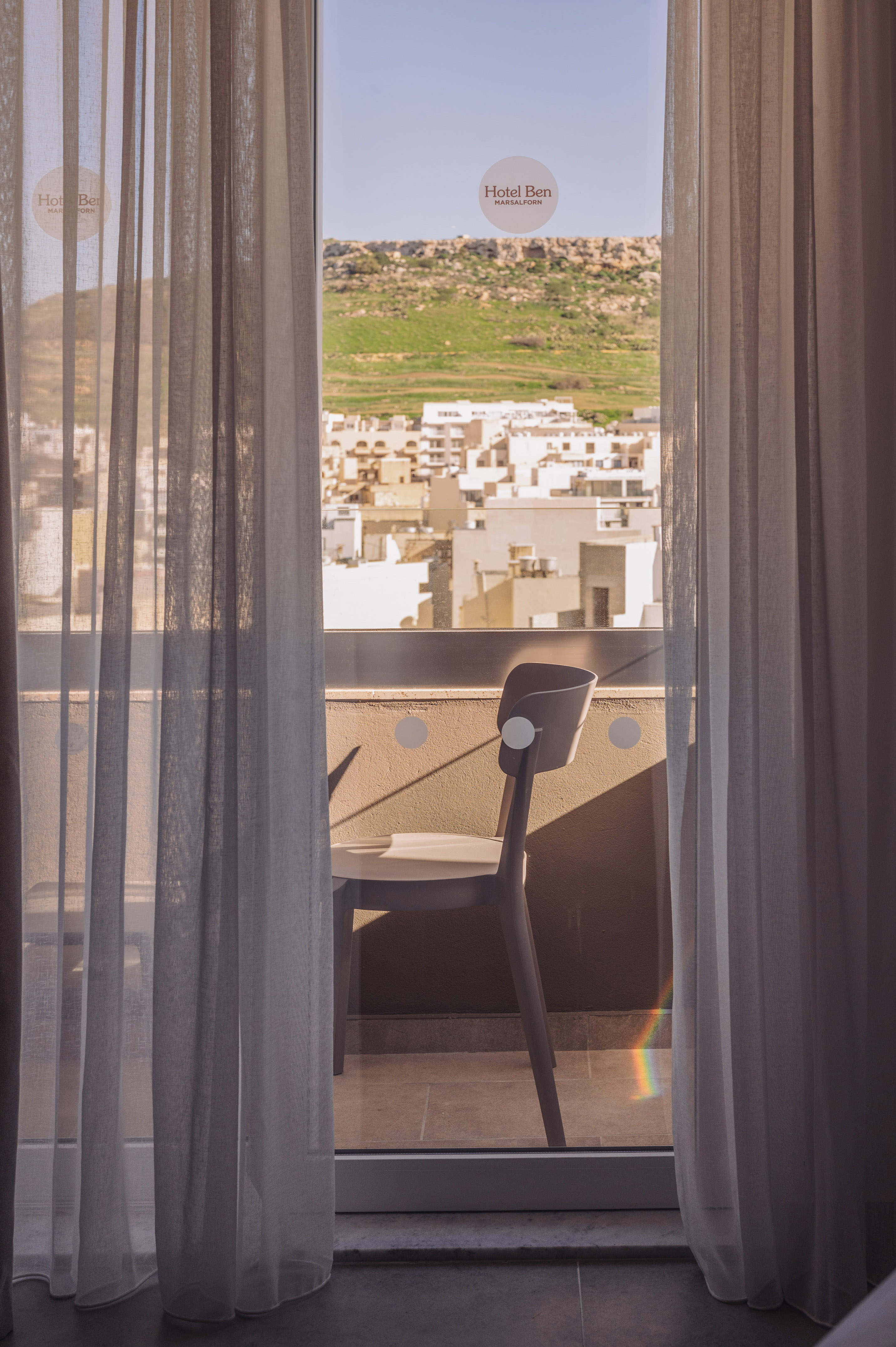 Hotel Ben Marsalforn room balcony overlooking cityscape & green hills.