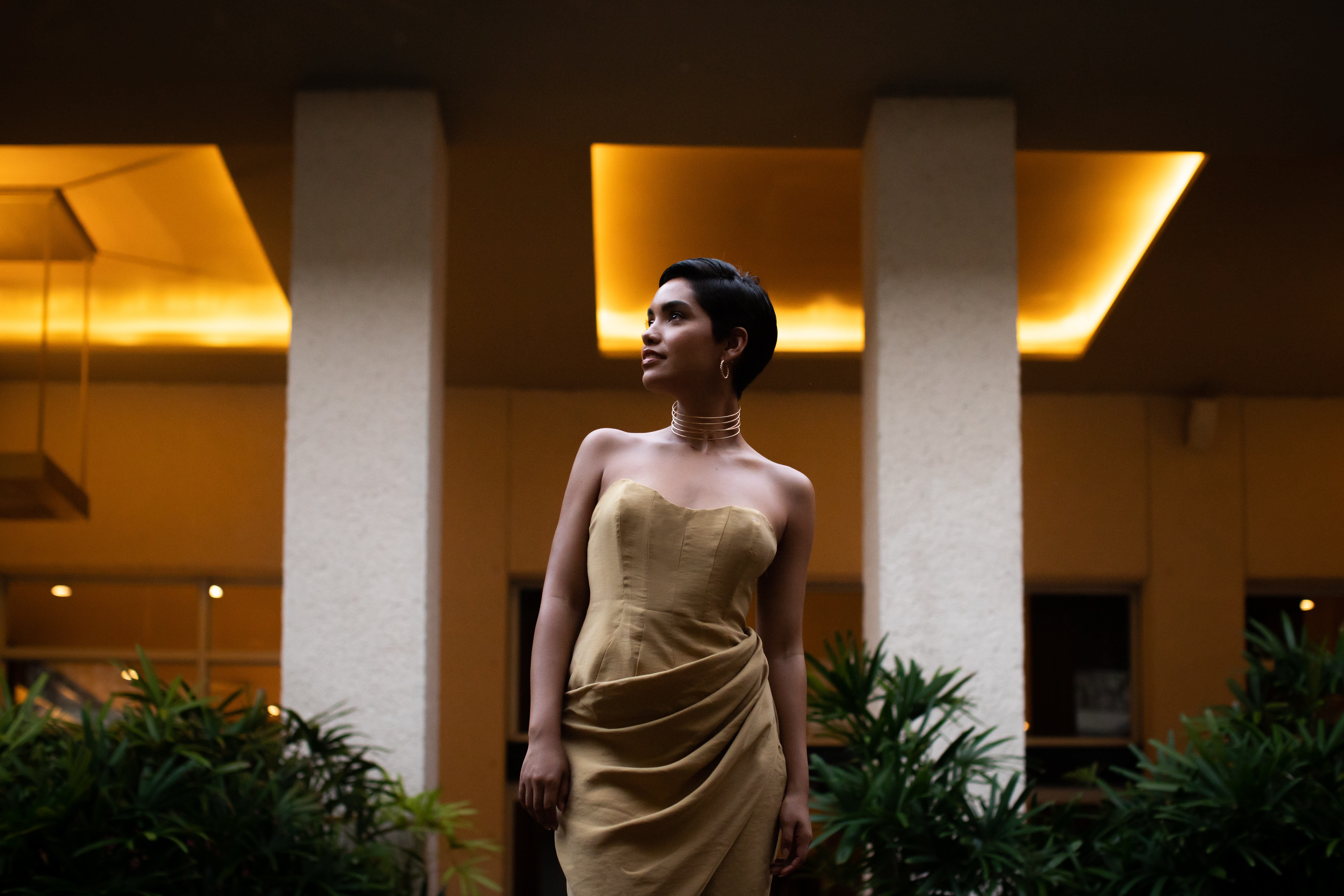 Woman in gold strapless dress, gold choker, short hair, modern building.