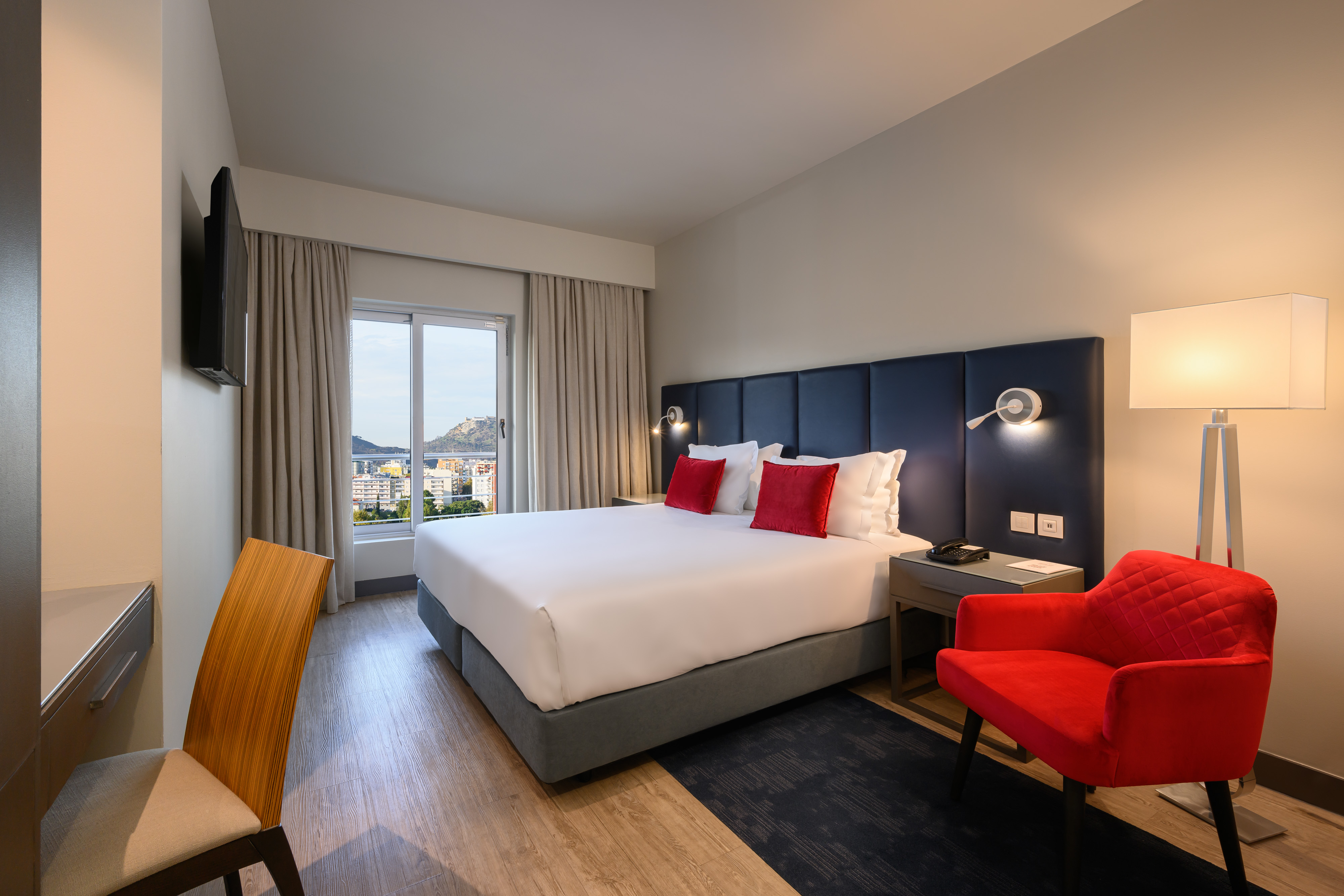 Modern hotel room with king bed, red armchair, city view, and desk.