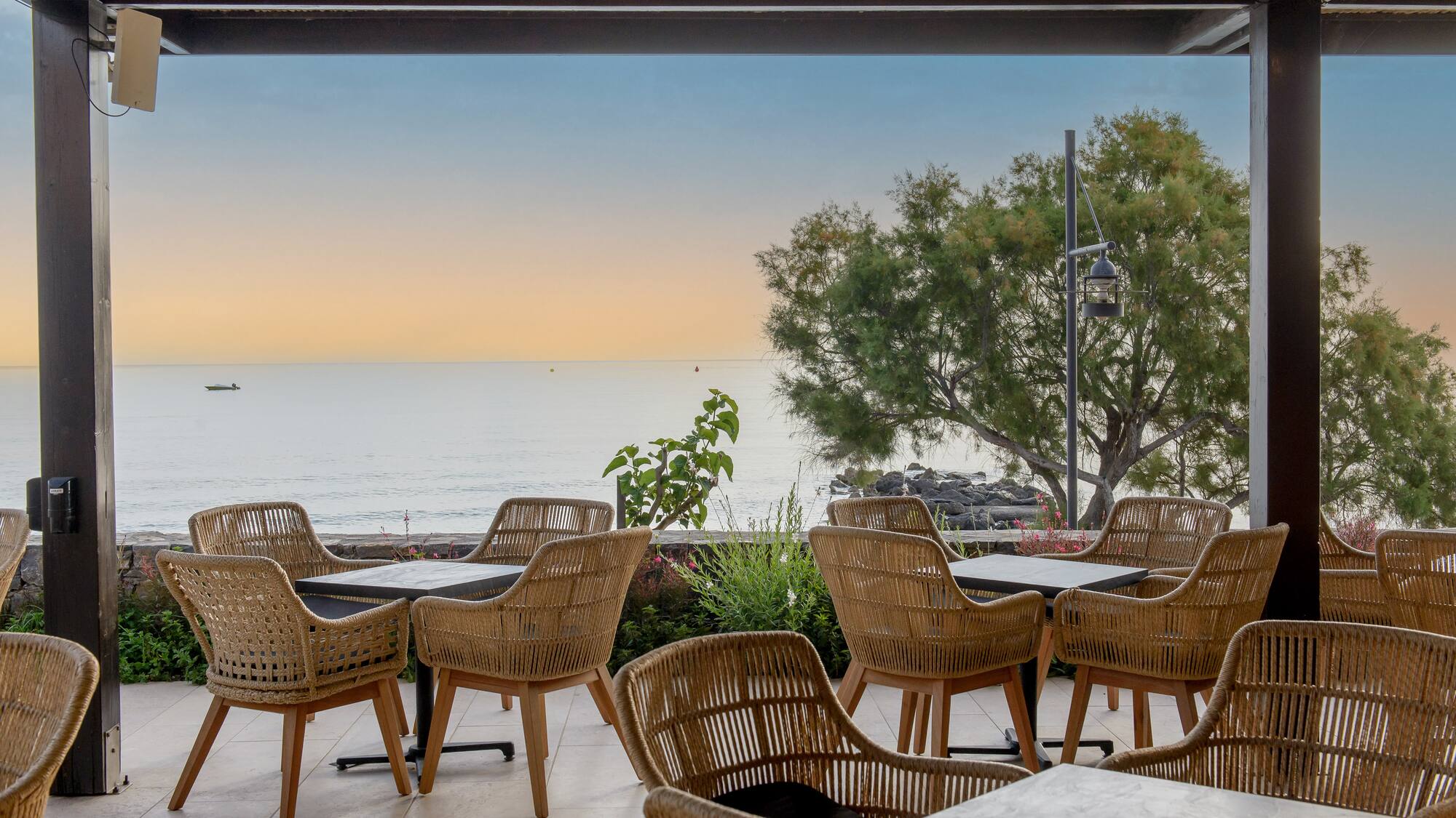 Waterfront dining patio with wicker chairs, ocean view at sunset.