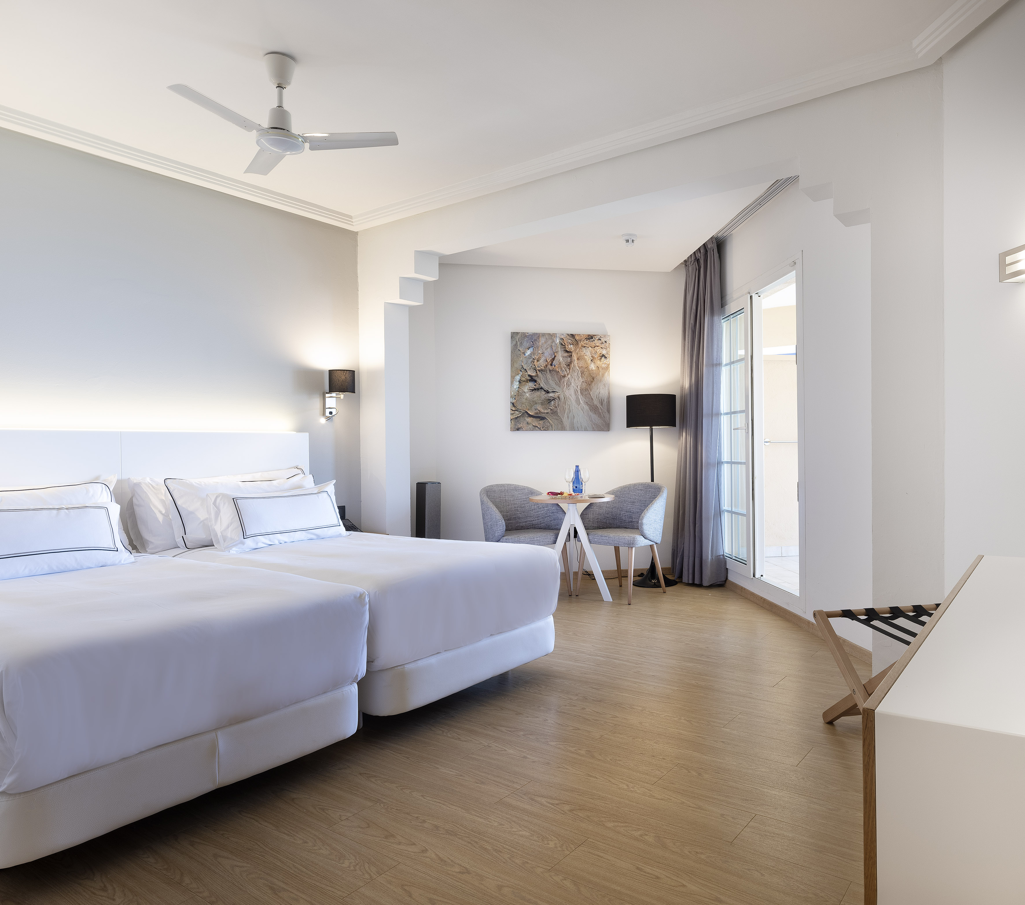 Bright modern hotel room with twin beds, ceiling fan, wood floor, balcony.