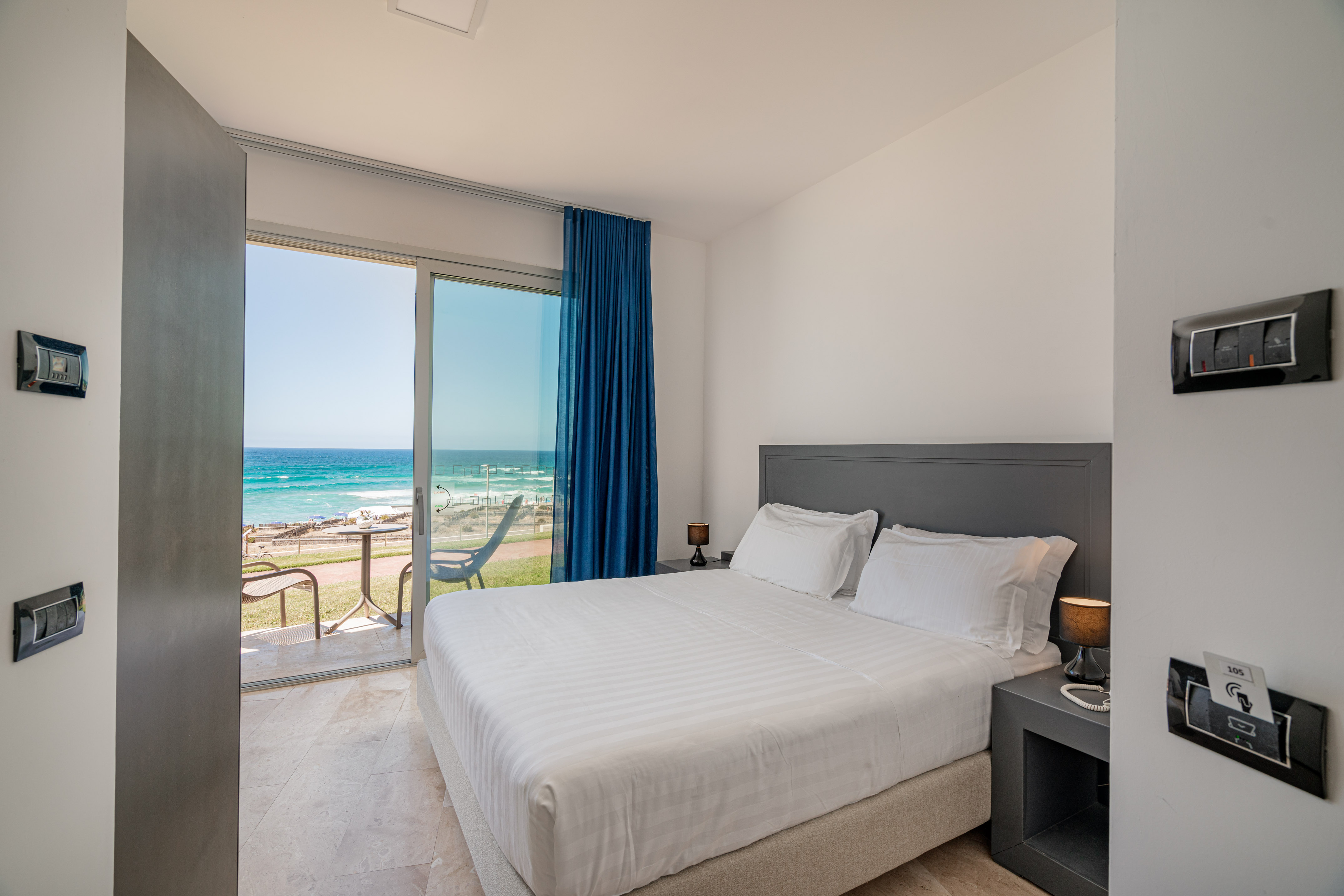 Modern hotel bedroom with white bed, sea view balcony, and blue curtains.