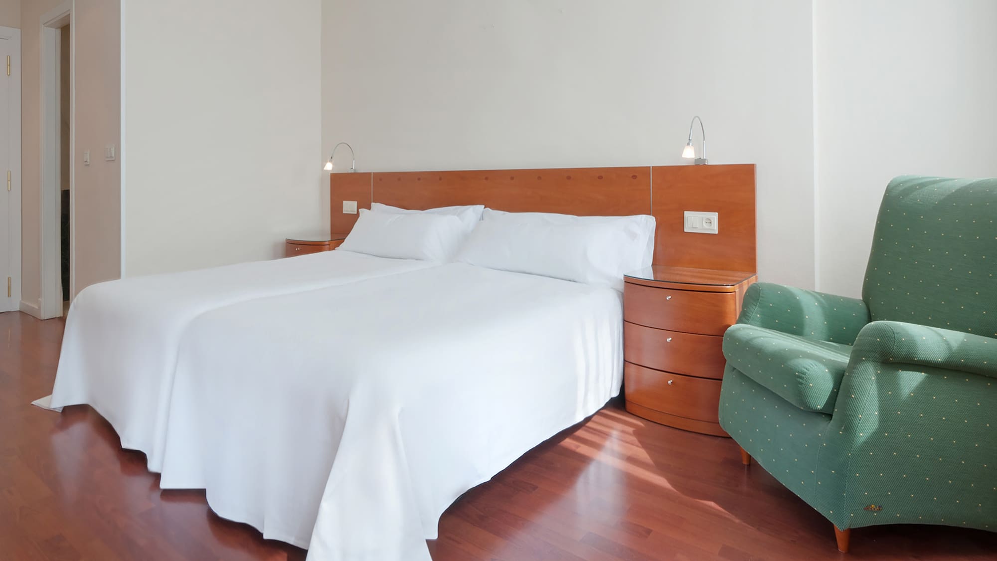 Hotel room with twin beds, white bedding, wood nightstands, and green chair.