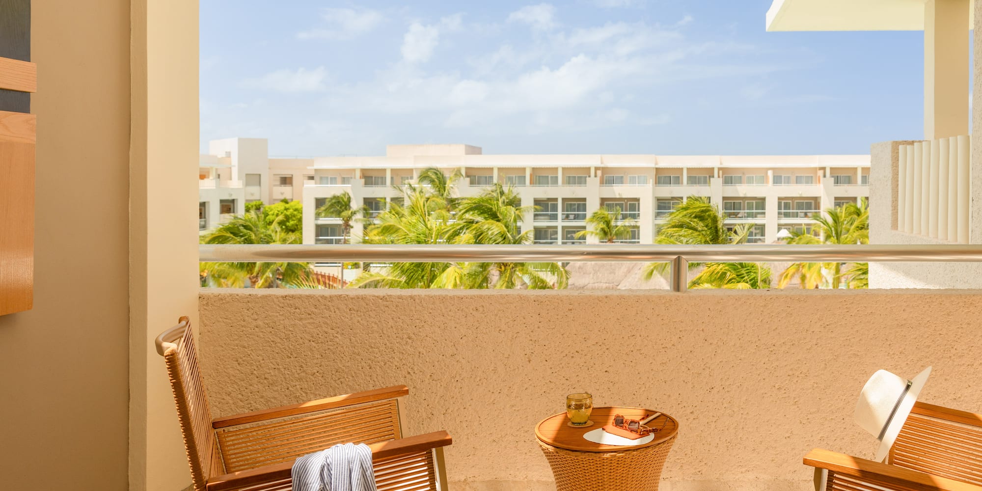 Sunny resort balcony with chairs, table, and palm tree hotel view.