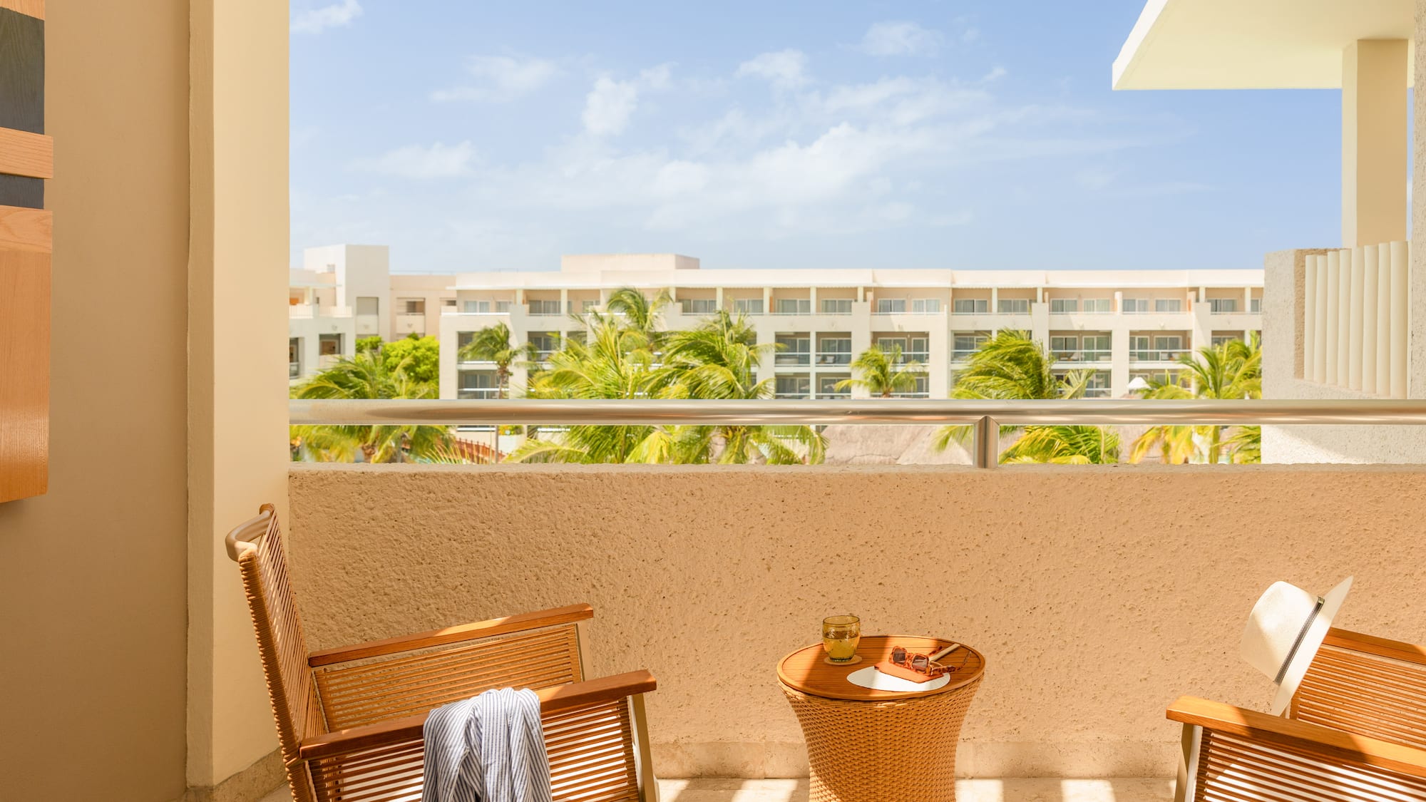 Sunny resort balcony with chairs, table, and palm tree hotel view.