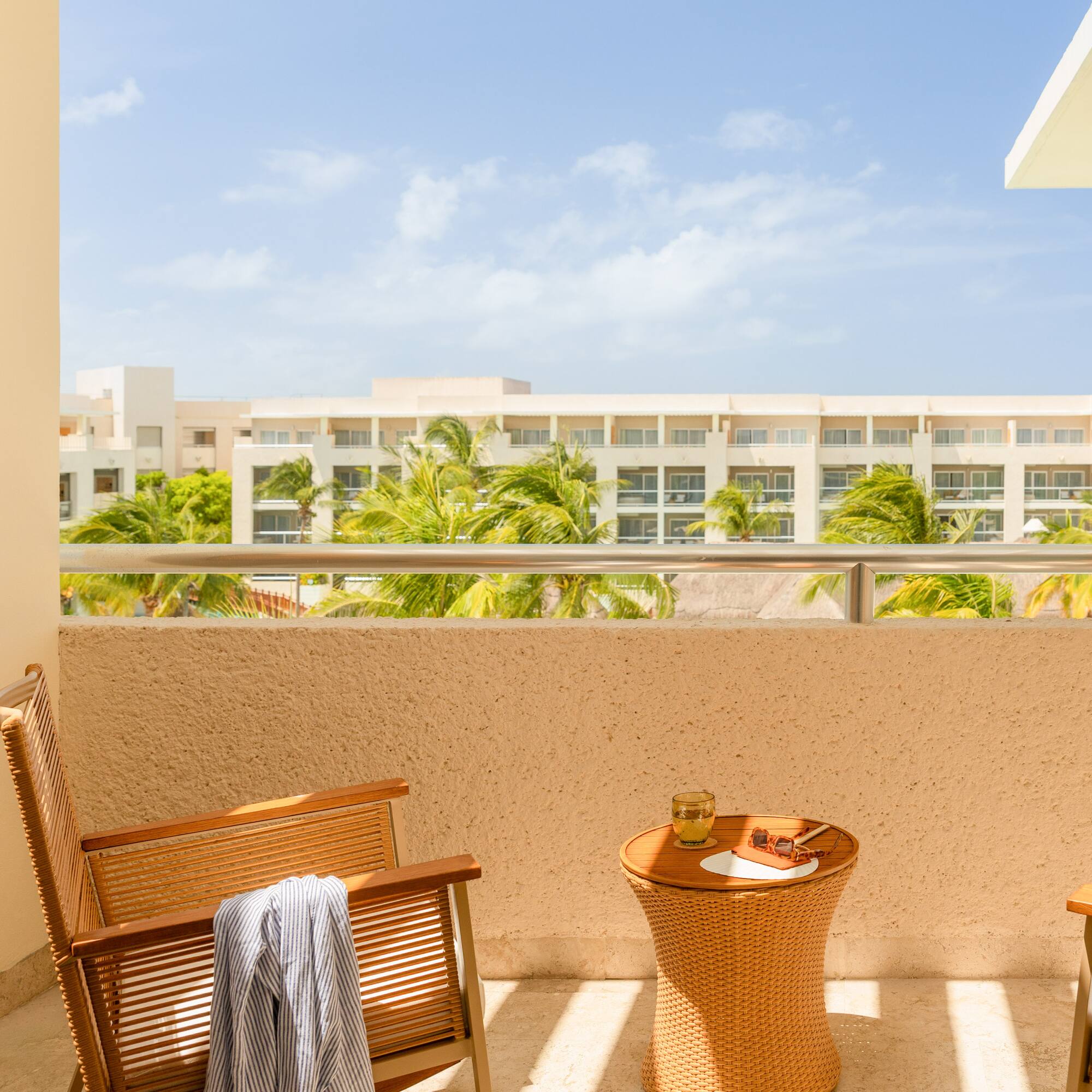 Sunny resort balcony with chairs, table, and palm tree hotel view.