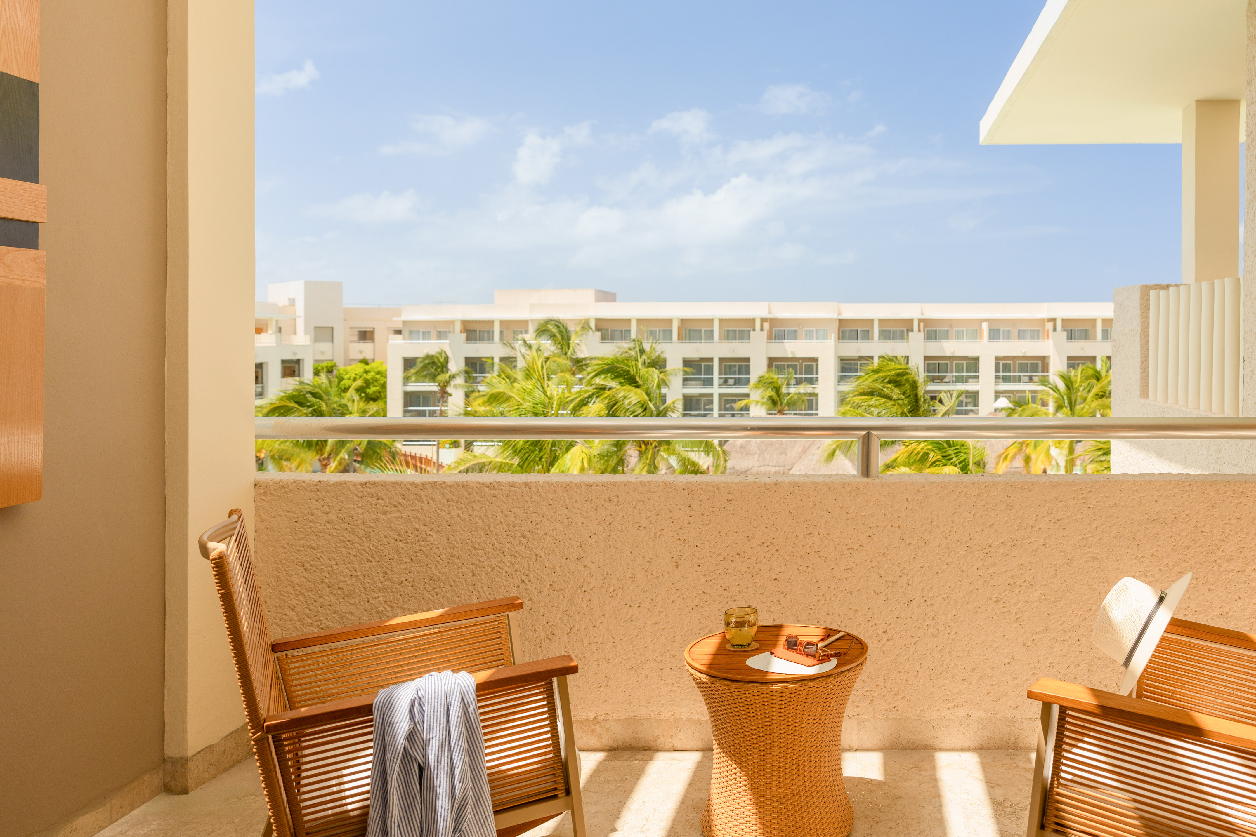 Sunny resort balcony with chairs, table, and palm tree hotel view.