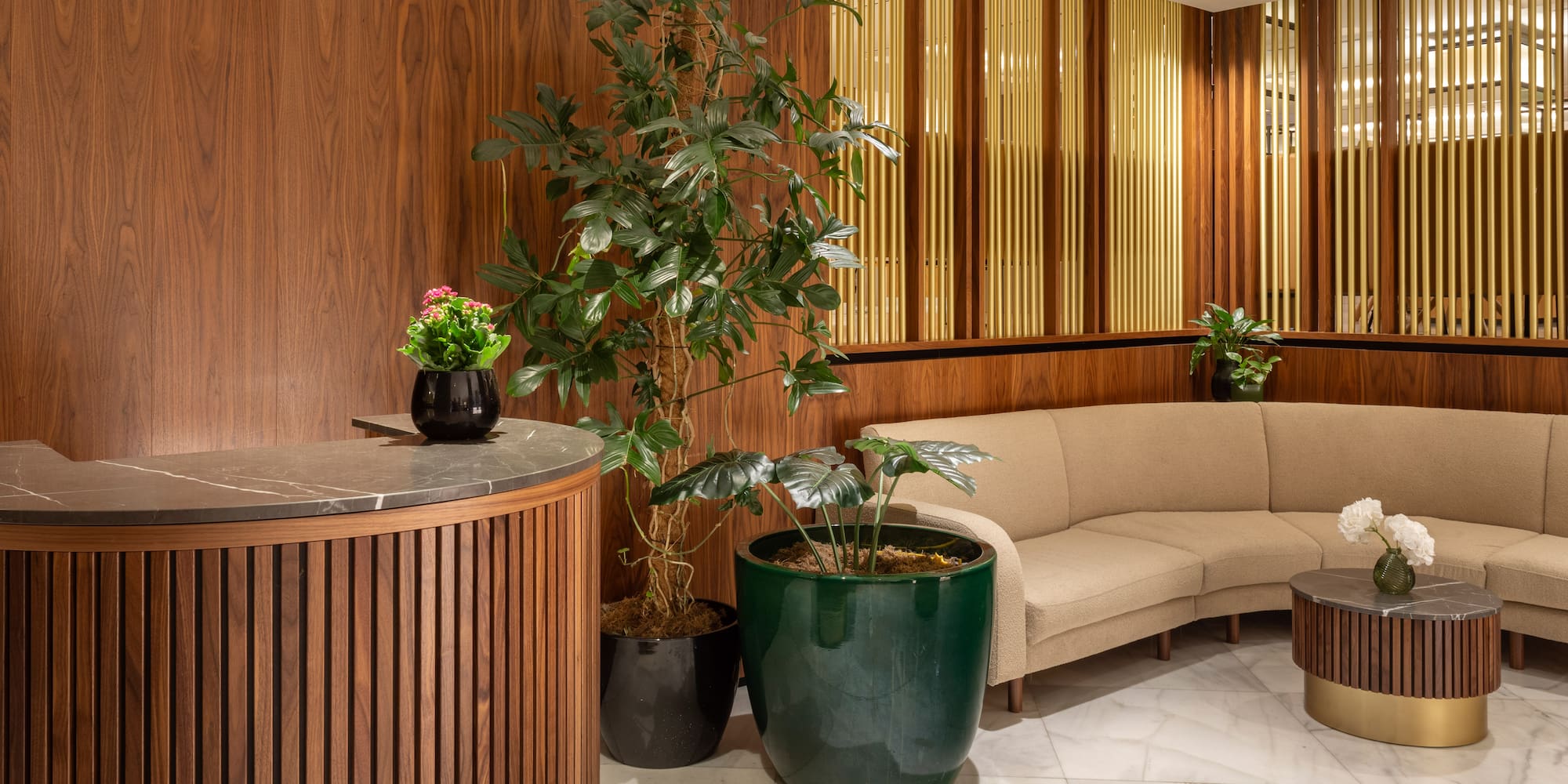 Modern lobby with wood reception, plants, curved sofa, marble, and gold accents.