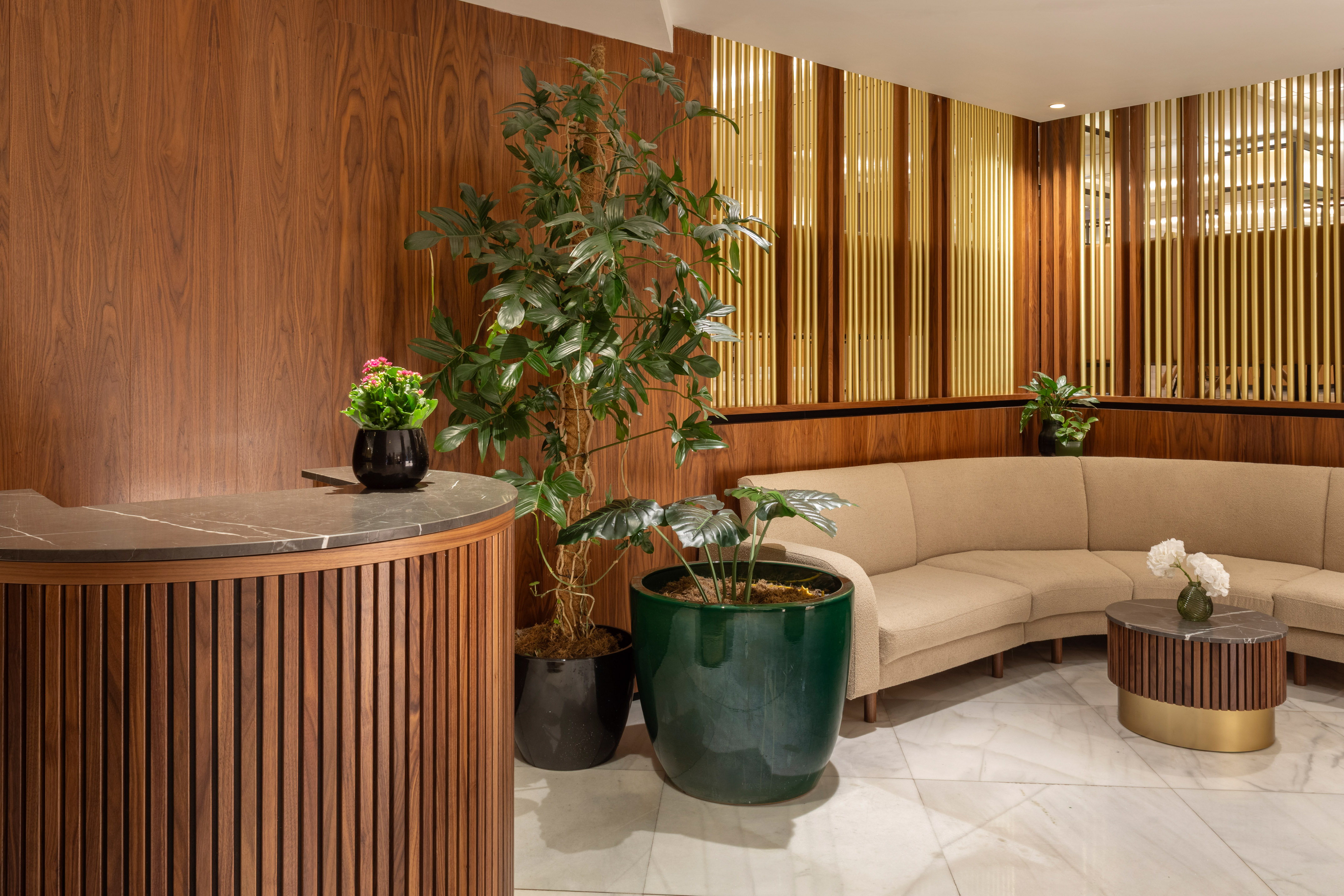 Modern lobby with wood reception, plants, curved sofa, marble, and gold accents.