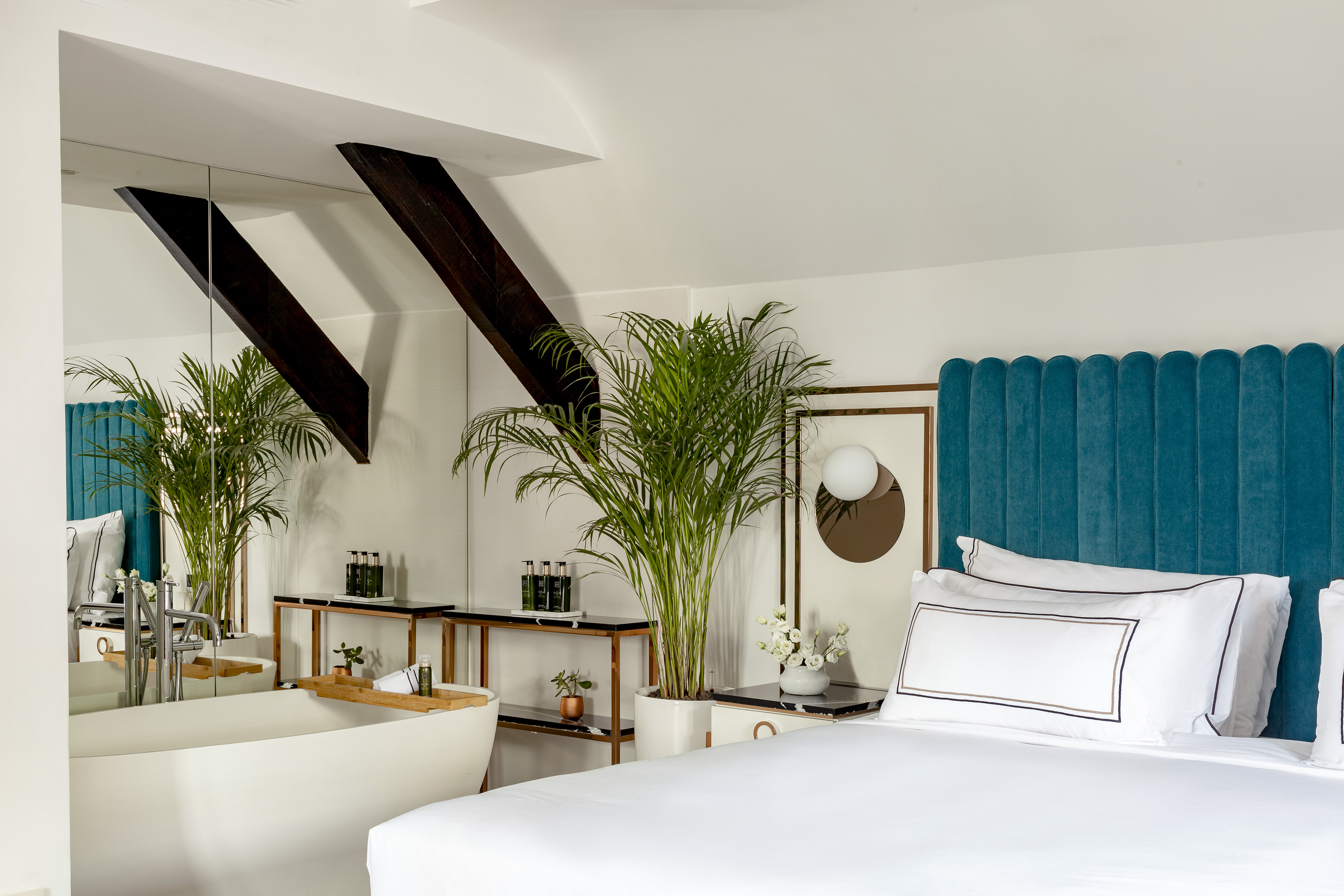Modern luxury hotel bedroom with teal headboard, white bed, and freestanding bathtub.