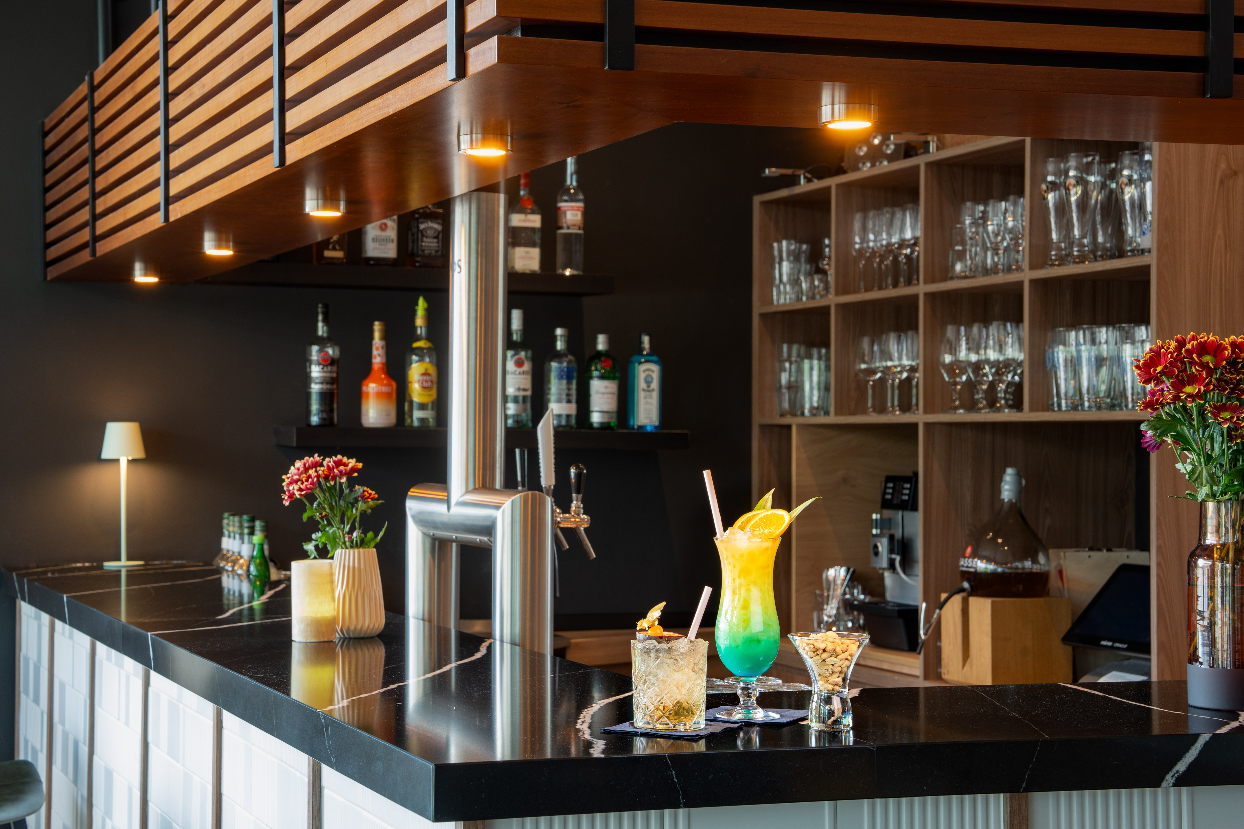 Modern bar counter with two colorful cocktails, liquor bottles, and wood details.