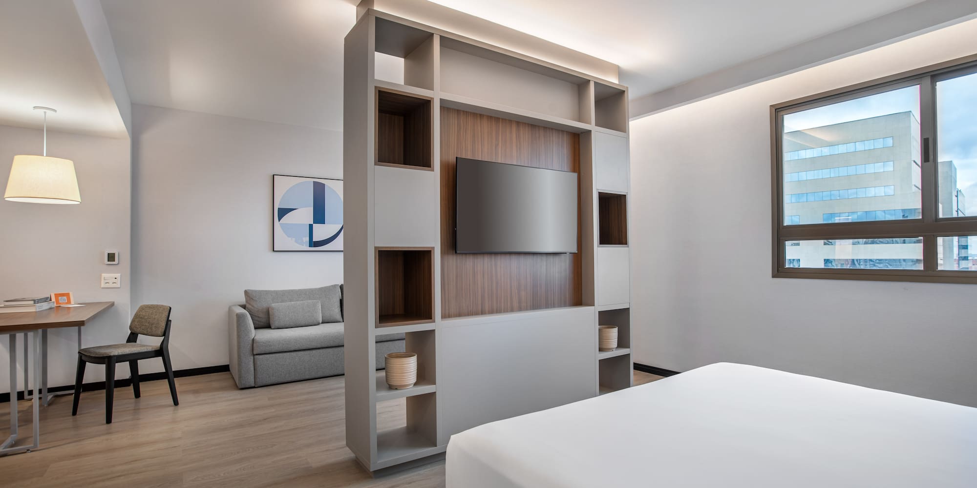 Modern hotel studio with bed, TV divider, sofa, and workspace.