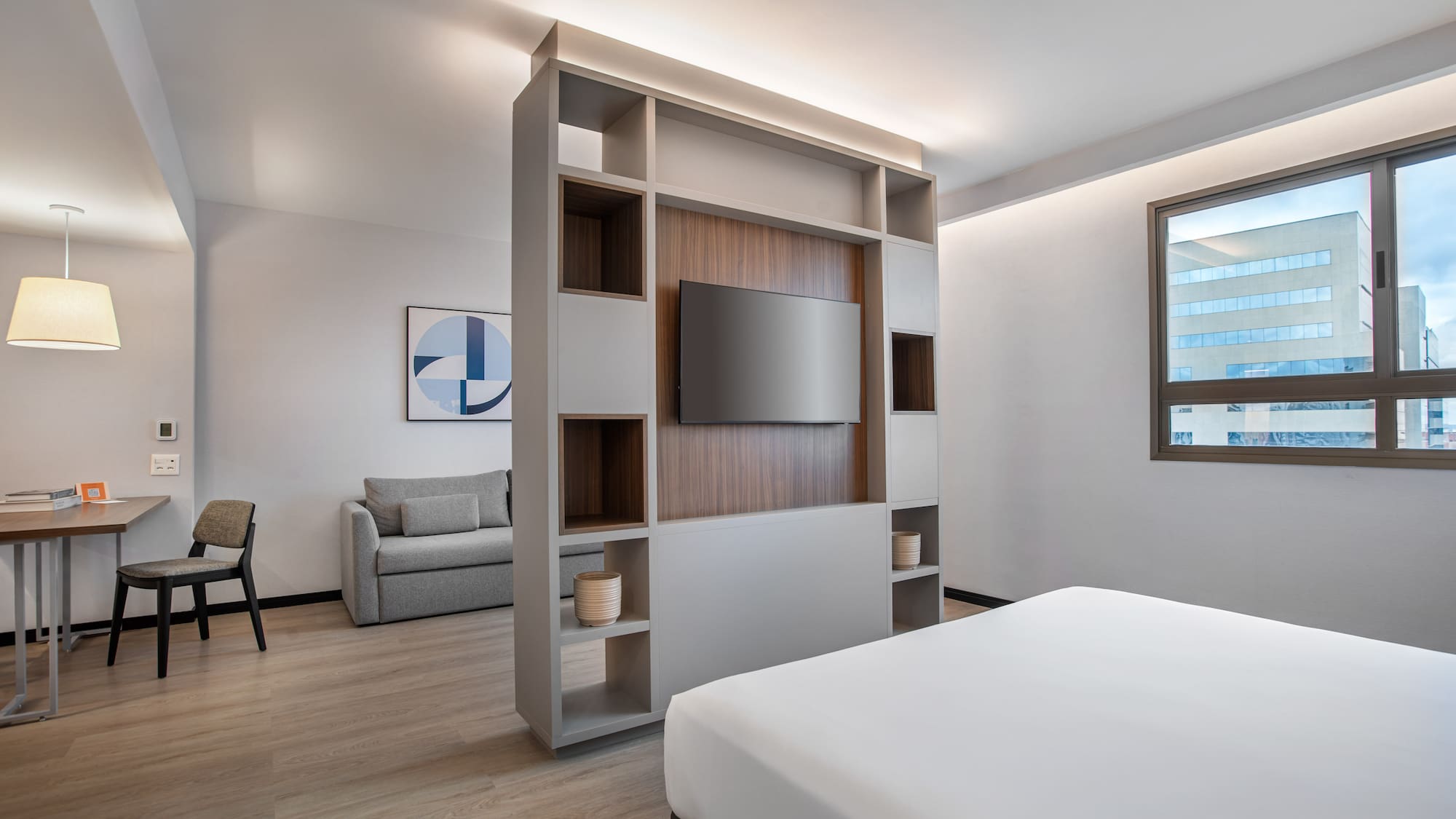 Modern hotel studio with bed, TV divider, sofa, and workspace.