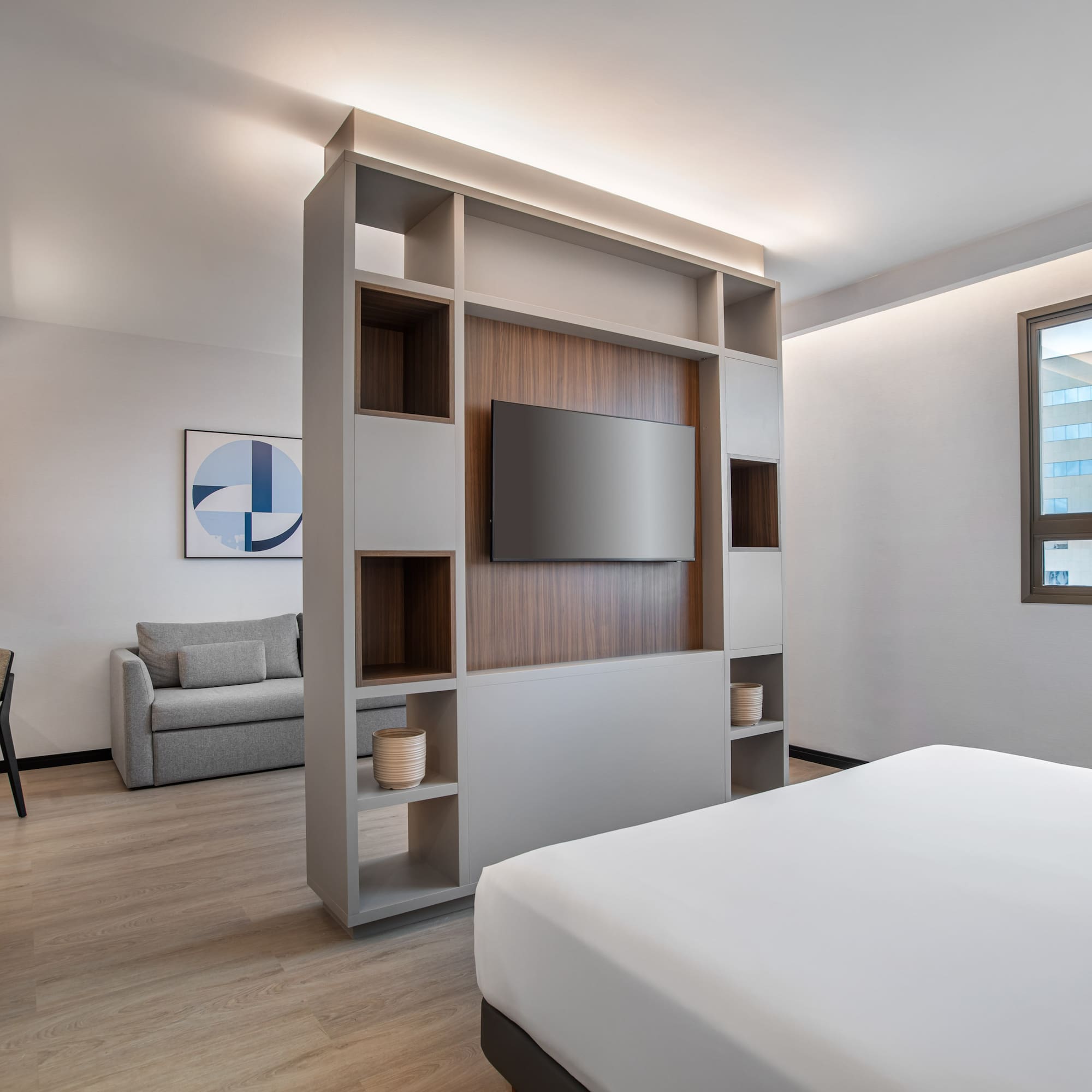 Modern hotel studio with bed, TV divider, sofa, and workspace.