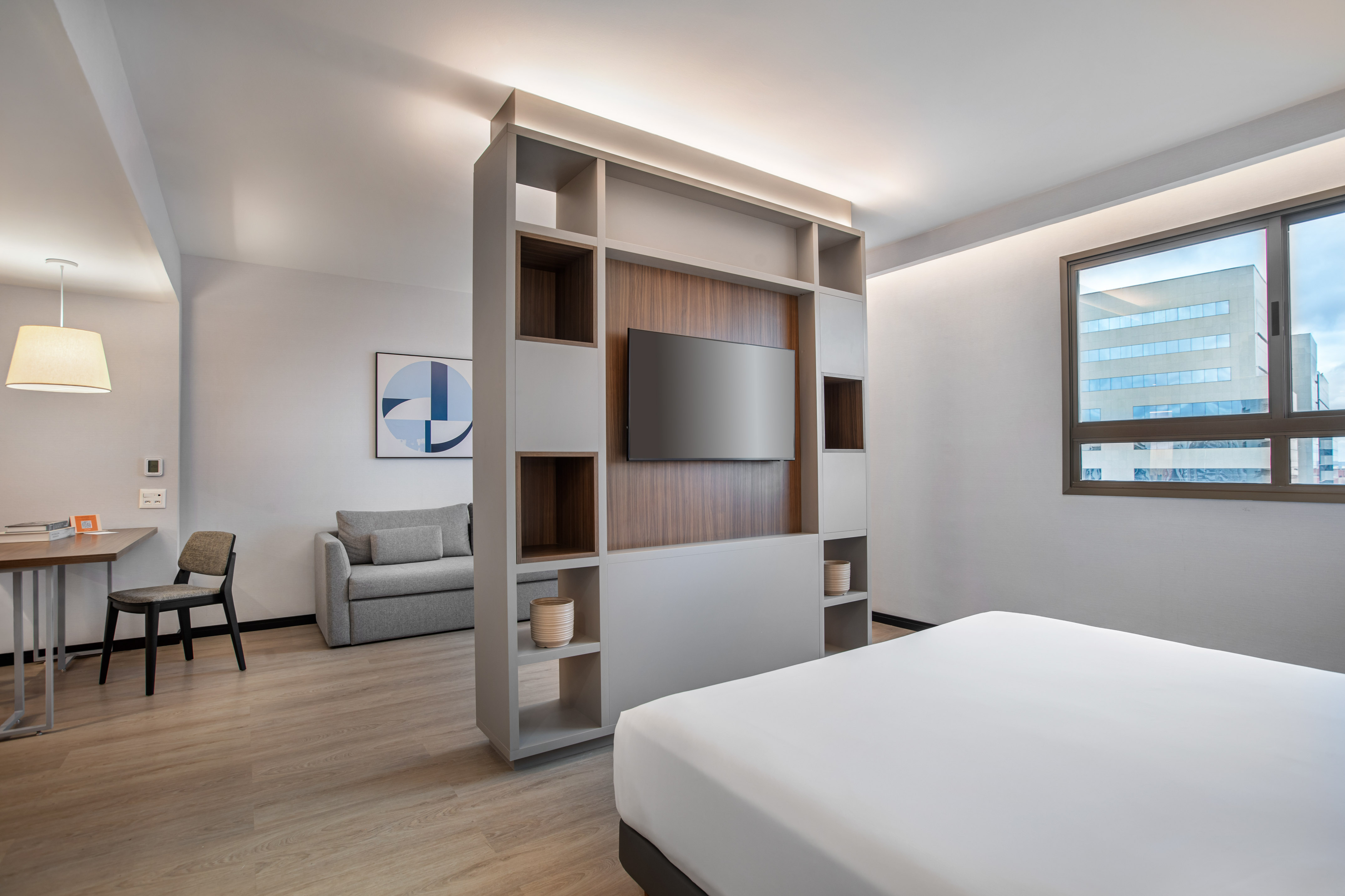 Modern hotel studio with bed, TV divider, sofa, and workspace.