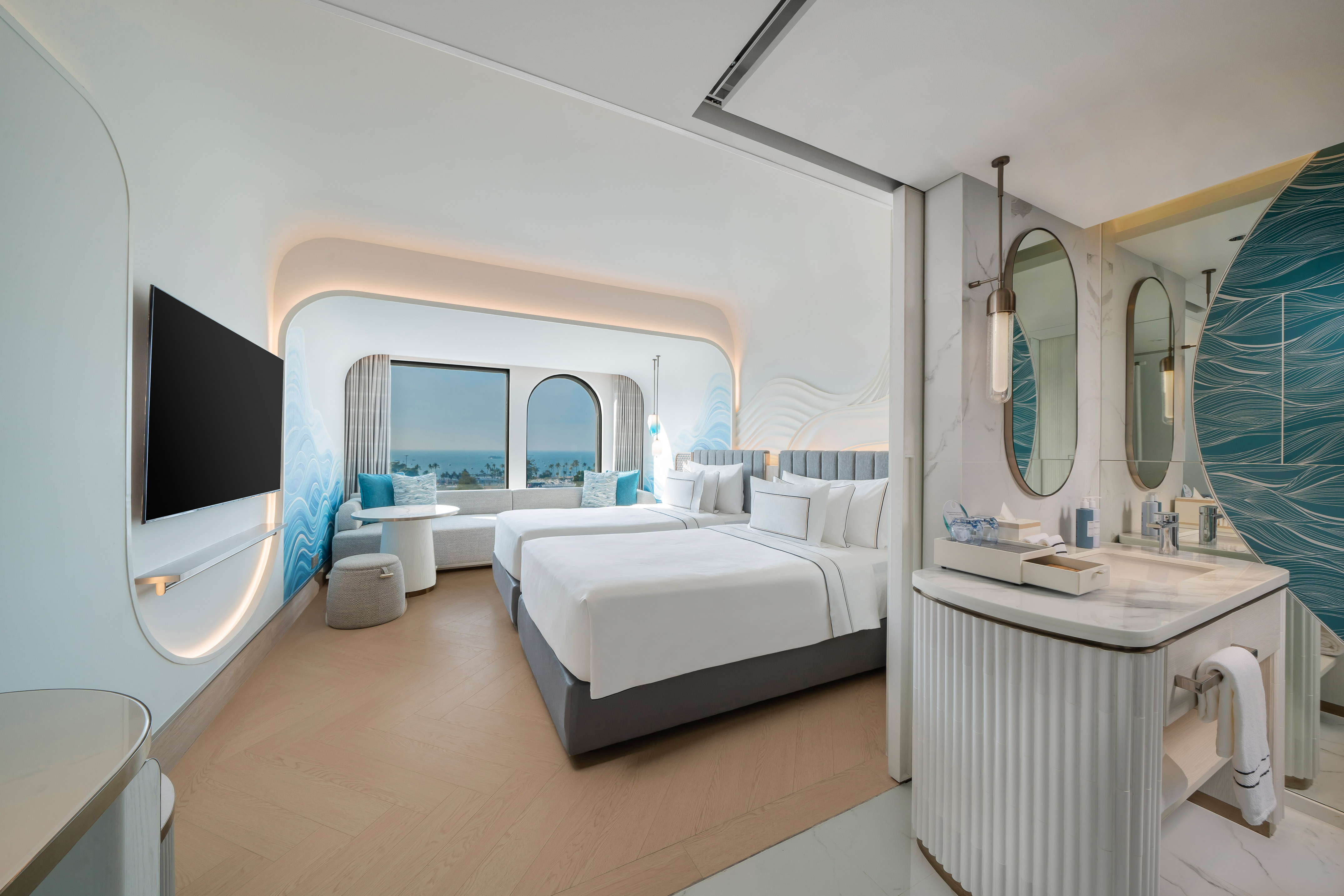 Modern hotel suite with ocean view, twin beds, blue wave decor, and vanity.