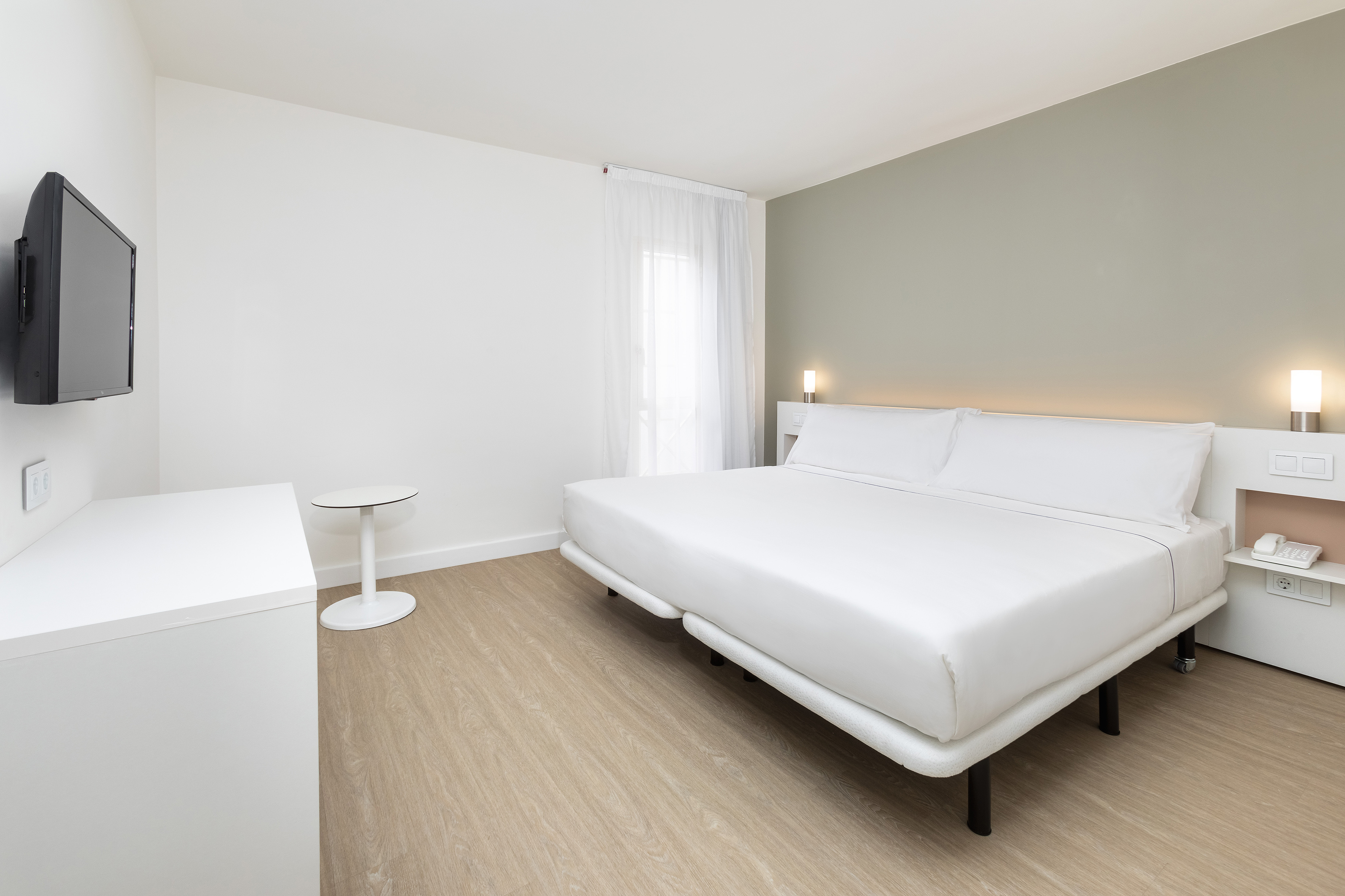 Modern minimalist hotel bedroom with large white bed, wall-mounted TV, light wood floor.