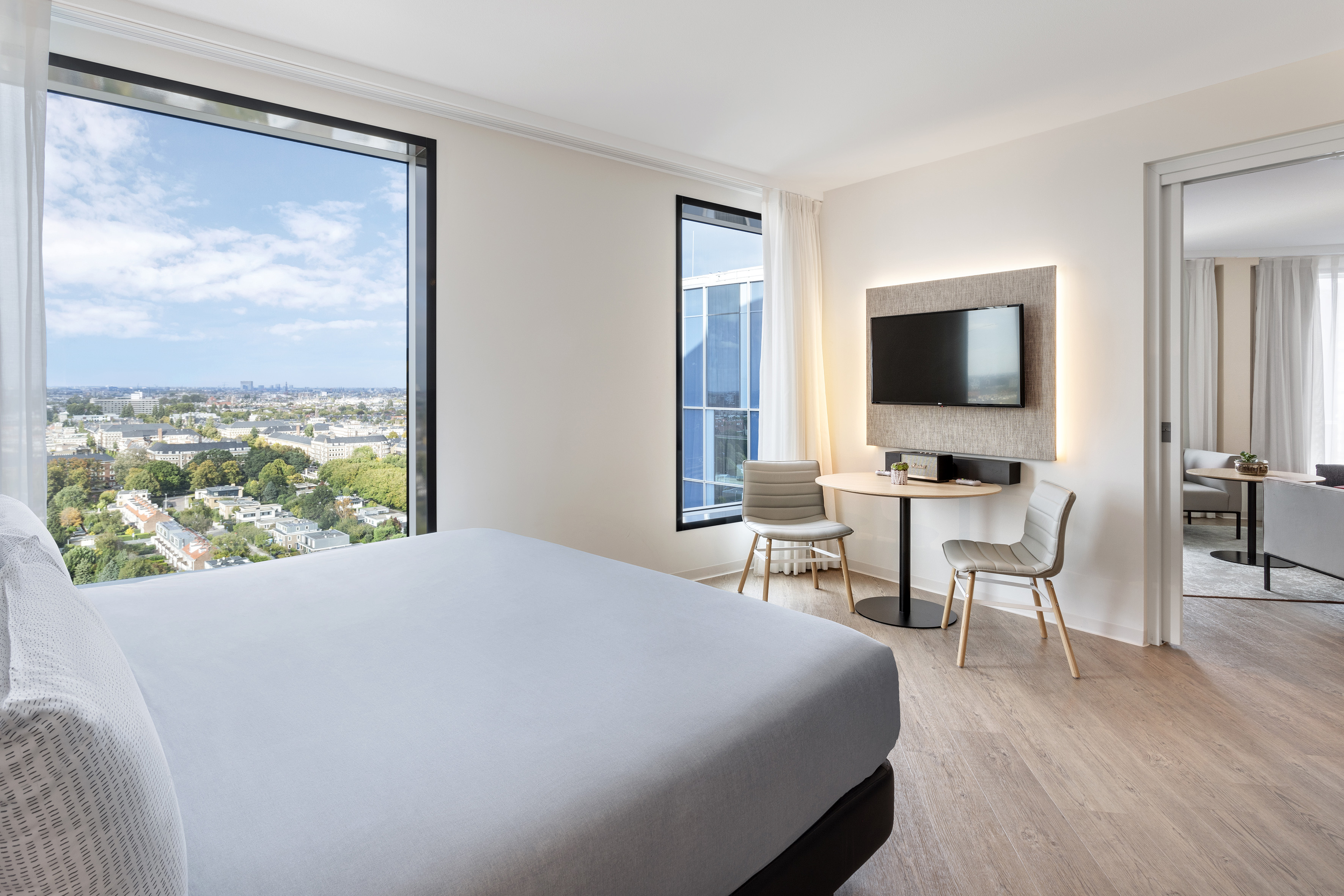 Modern hotel room with city view, large bed, flat-screen TV, and wood floor.