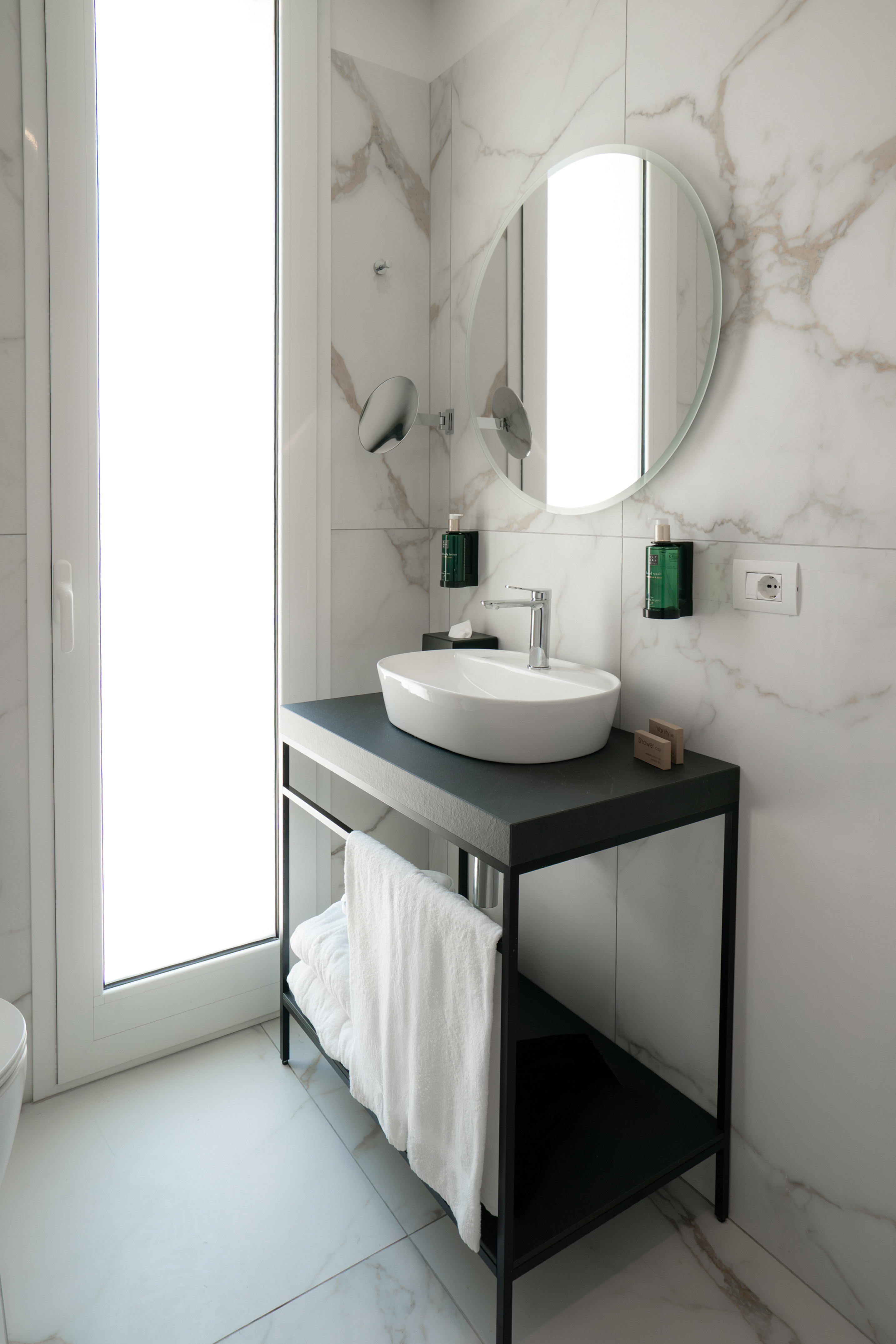 Modern bathroom: white marble walls, vessel sink, black vanity, round mirror.