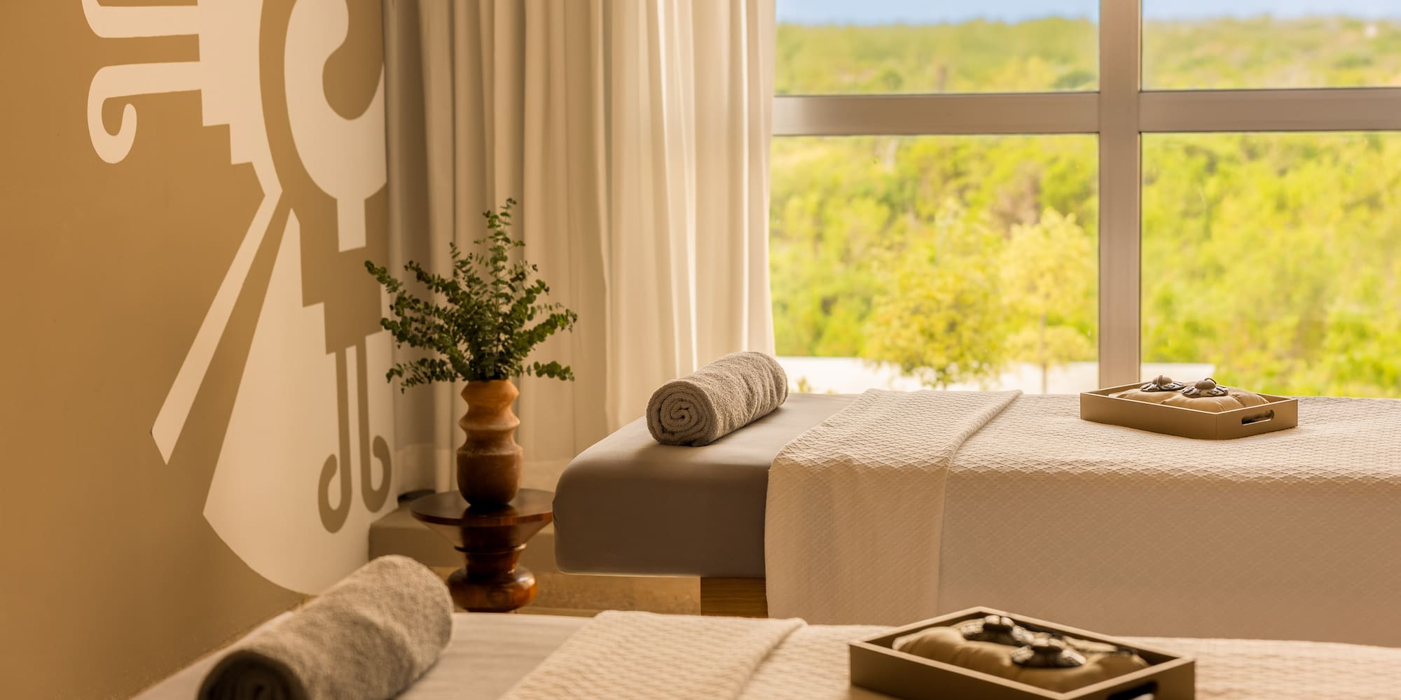 Serene spa with two massage beds, rolled towels, mural, and lush nature view.