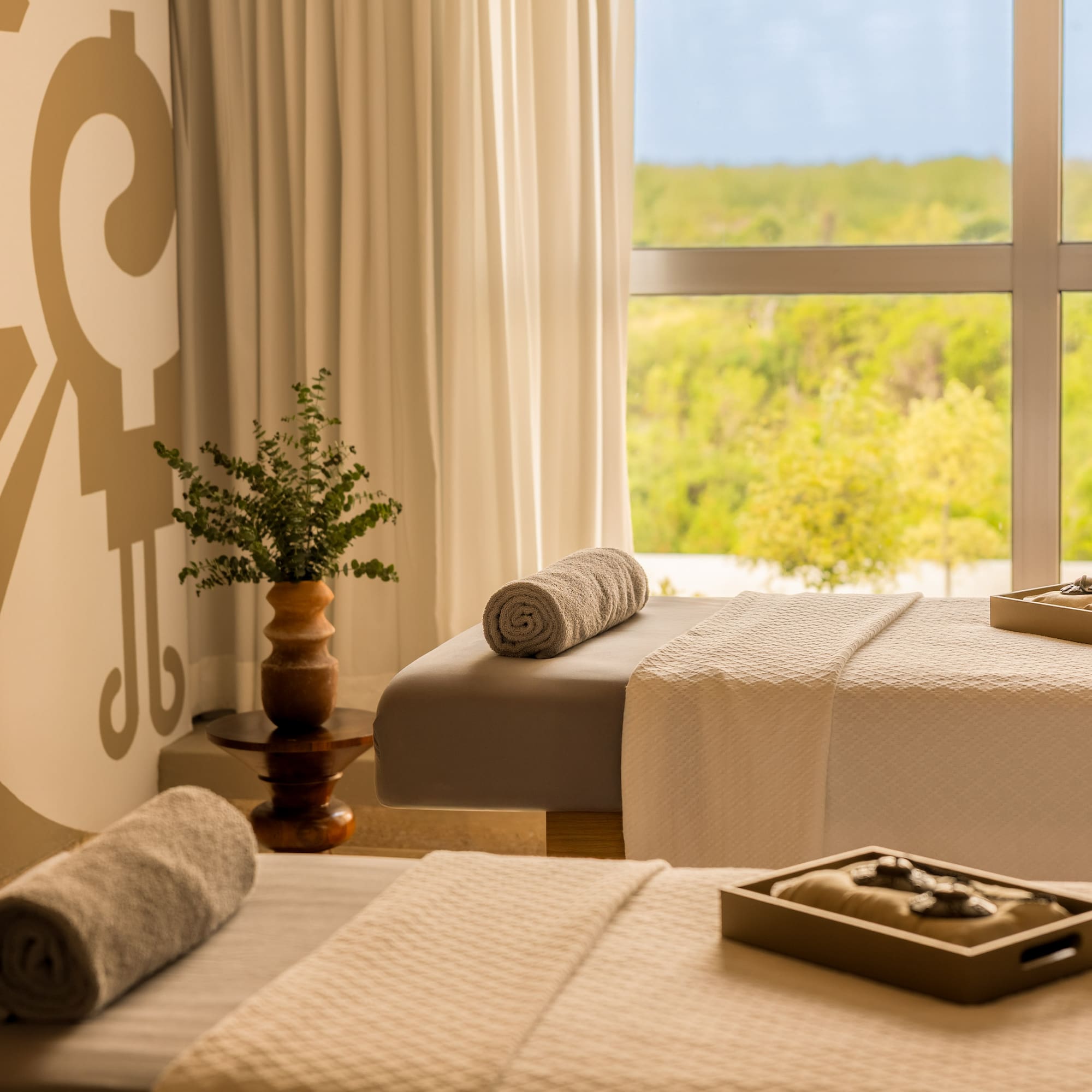 Serene spa with two massage beds, rolled towels, mural, and lush nature view.