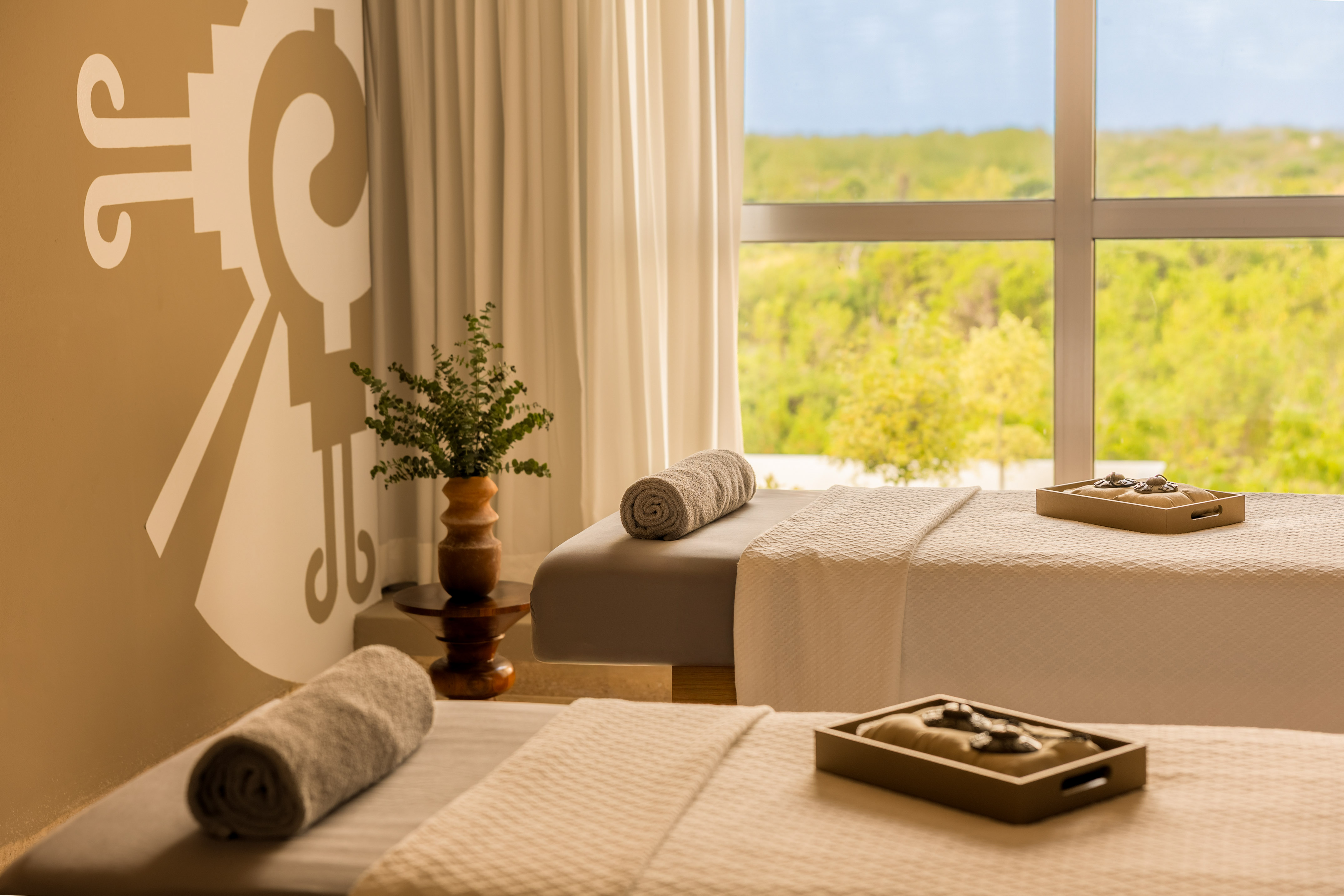 Serene spa with two massage beds, rolled towels, mural, and lush nature view.