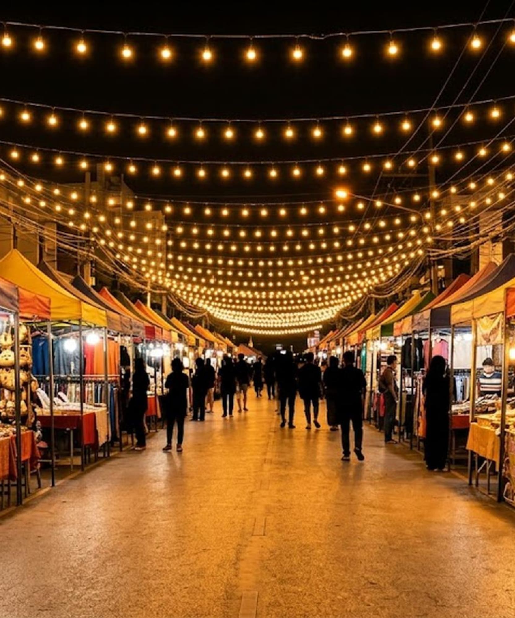 Night market street with colorful vendor stalls, string lights, and shoppers.