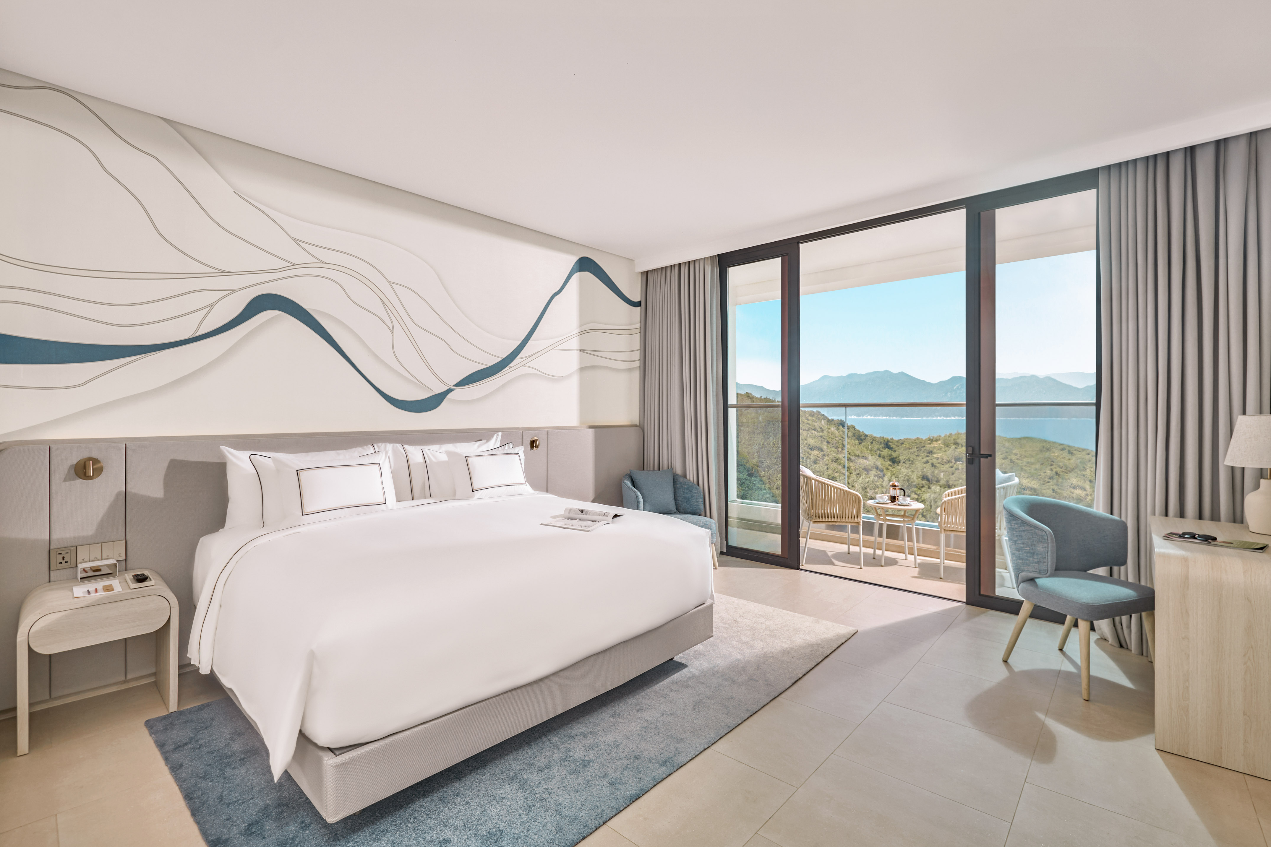 Modern hotel bedroom with ocean view balcony, white bed, blue decor.