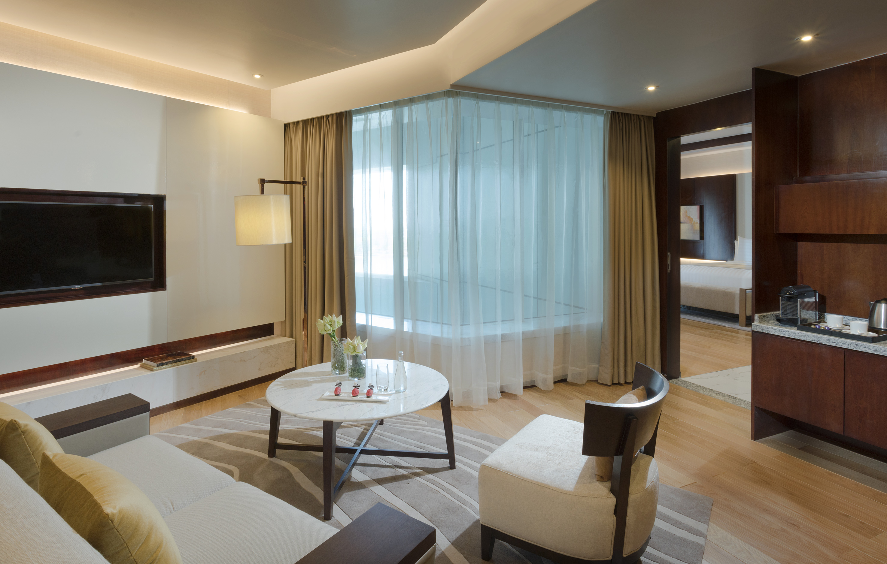 Modern hotel suite living room with sofa, TV, large window, and kitchenette.