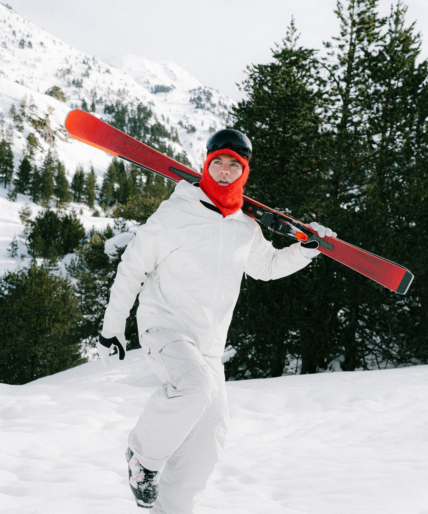 A skier in white winter gear carries skis in a snowy mountain landscape.