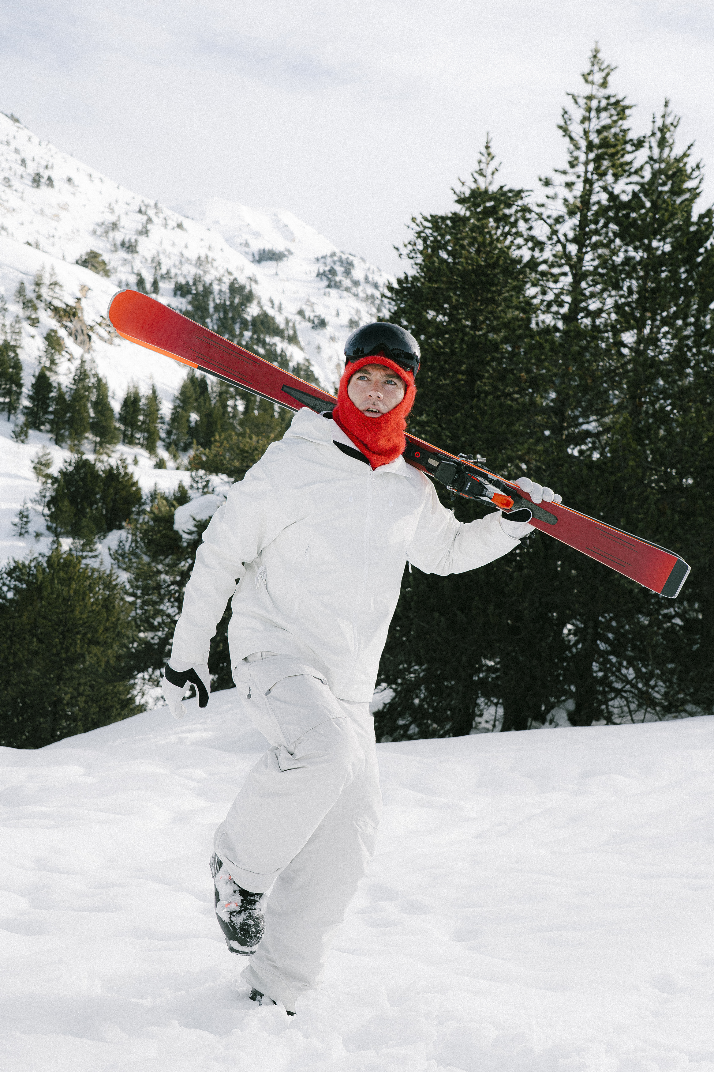 A skier in white winter gear carries skis in a snowy mountain landscape.