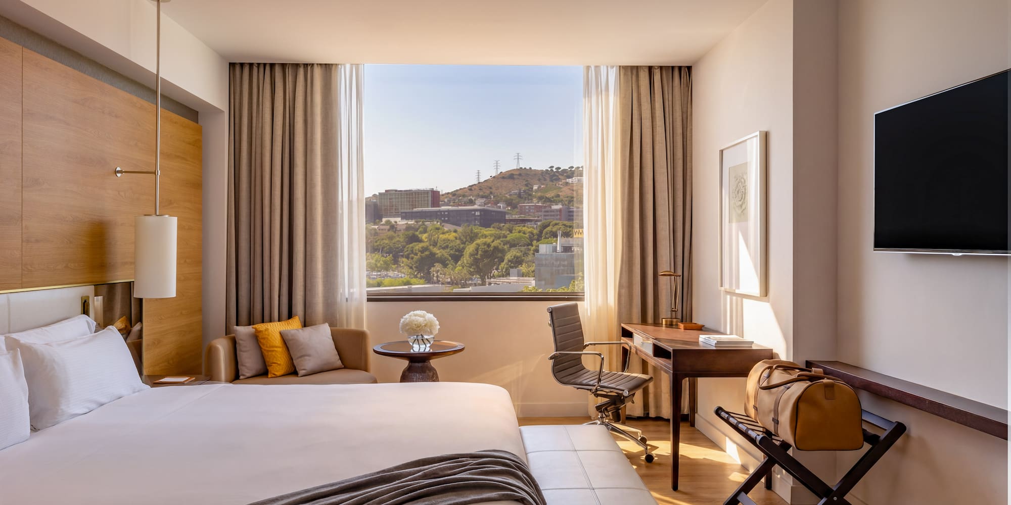Modern hotel room with city view, large bed, desk, and warm natural light.