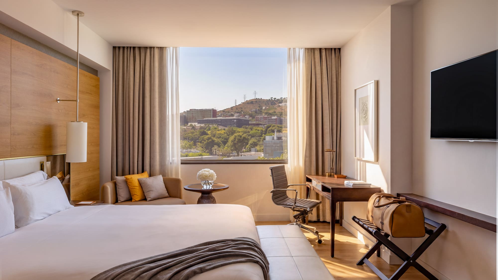 Modern hotel room with city view, large bed, desk, and warm natural light.