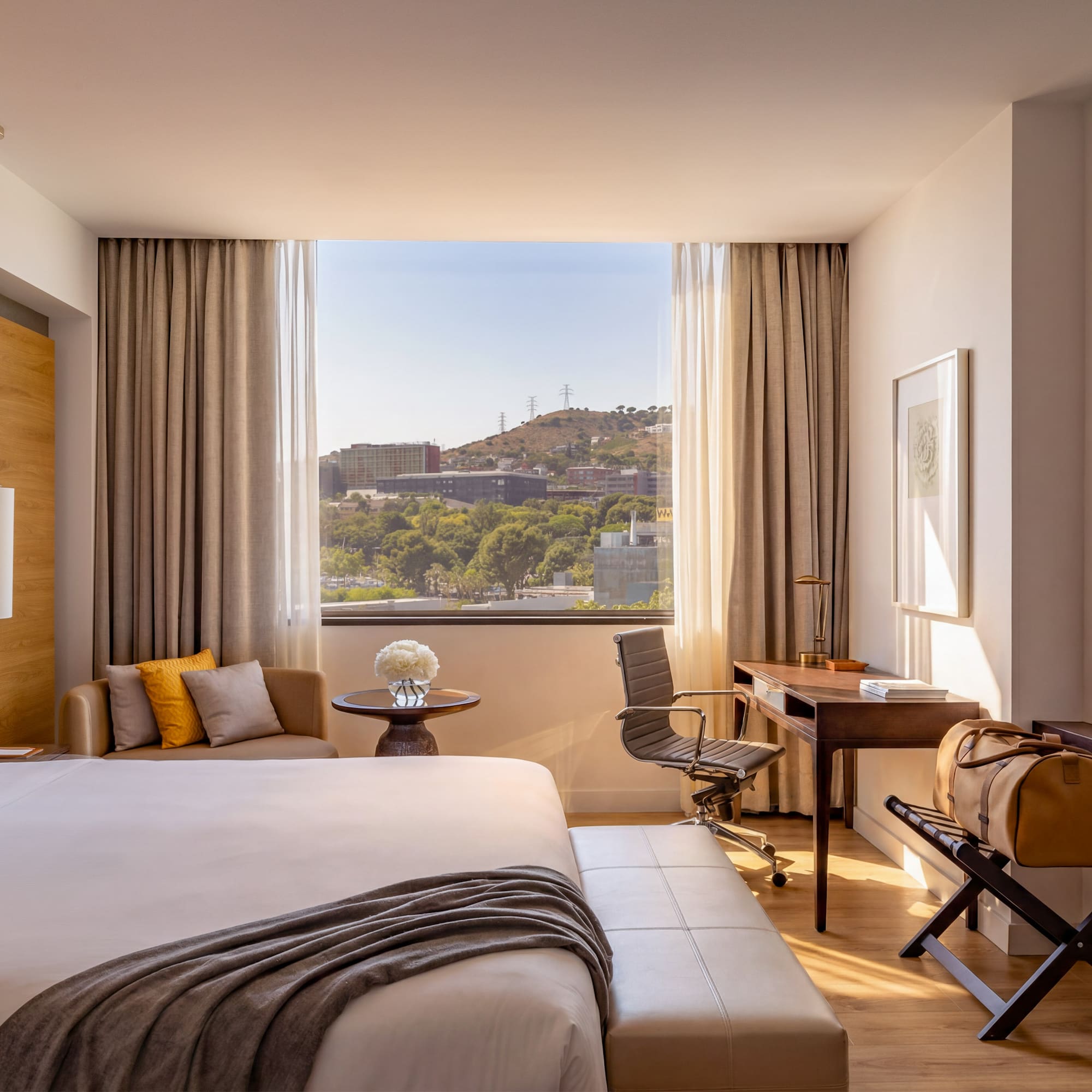 Modern hotel room with city view, large bed, desk, and warm natural light.