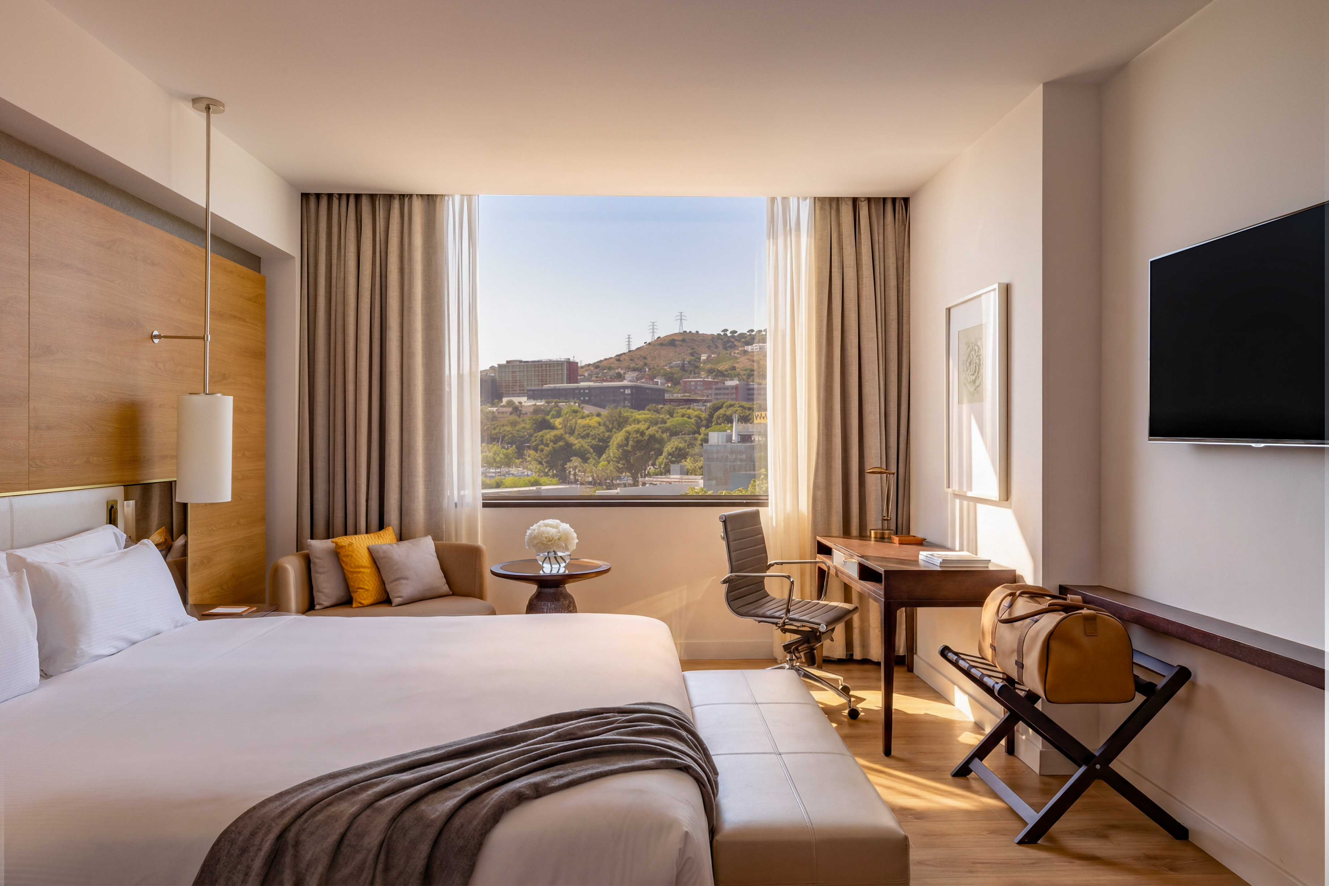 Modern hotel room with city view, large bed, desk, and warm natural light.