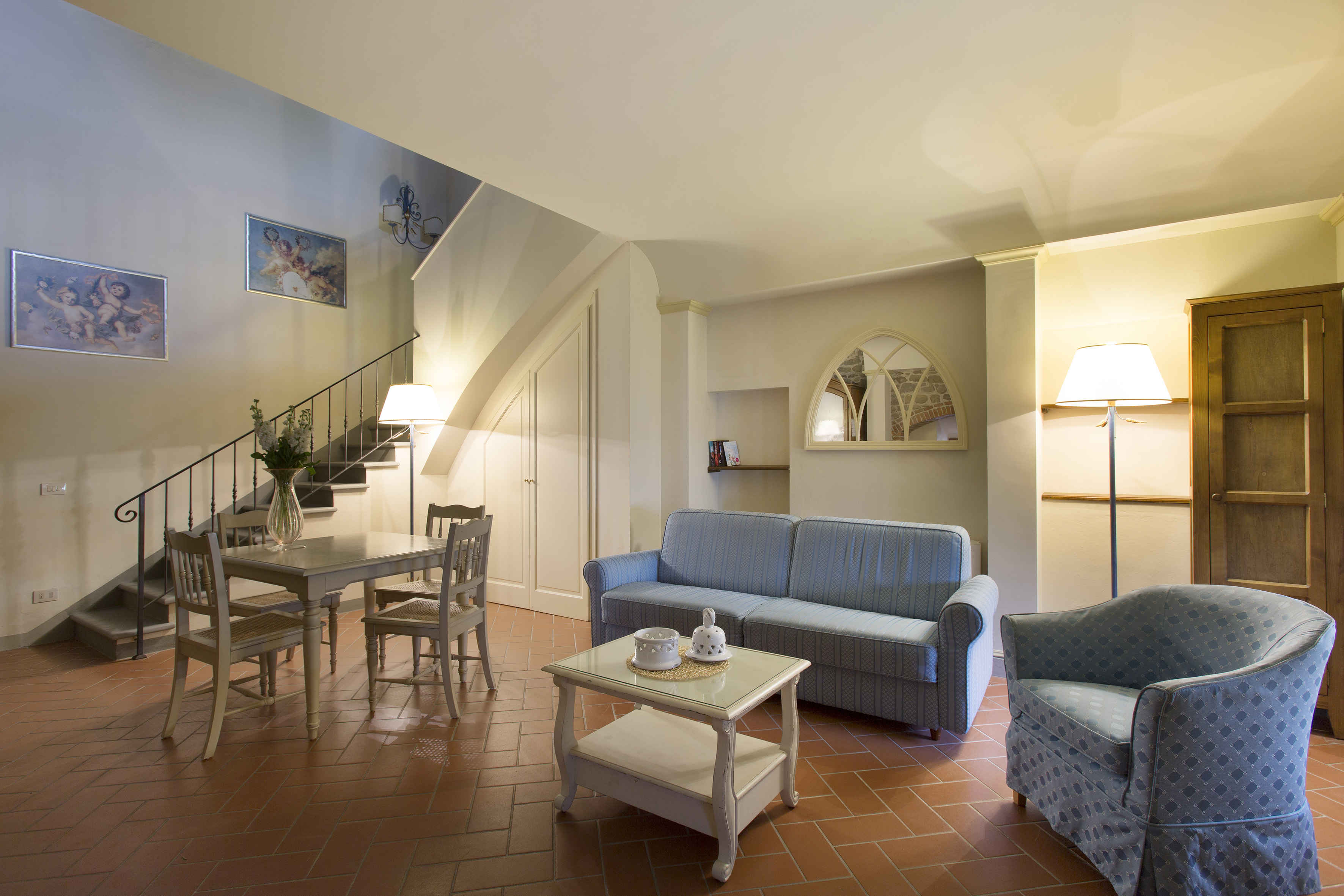 Cozy living room with blue sofa, dining area, terracotta floor, and staircase.