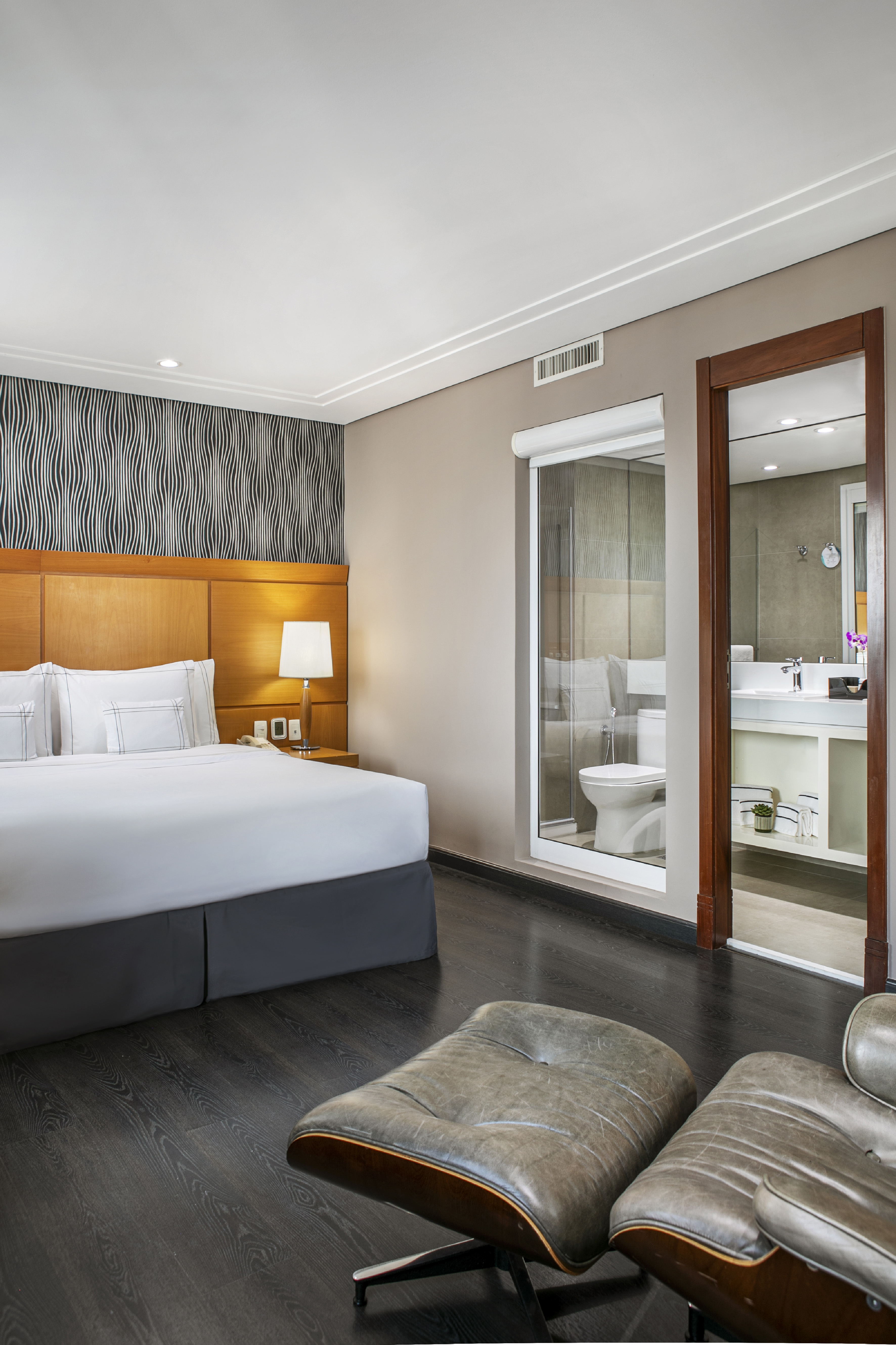Modern hotel room with king bed, glass-walled bathroom, and leather lounge chair.