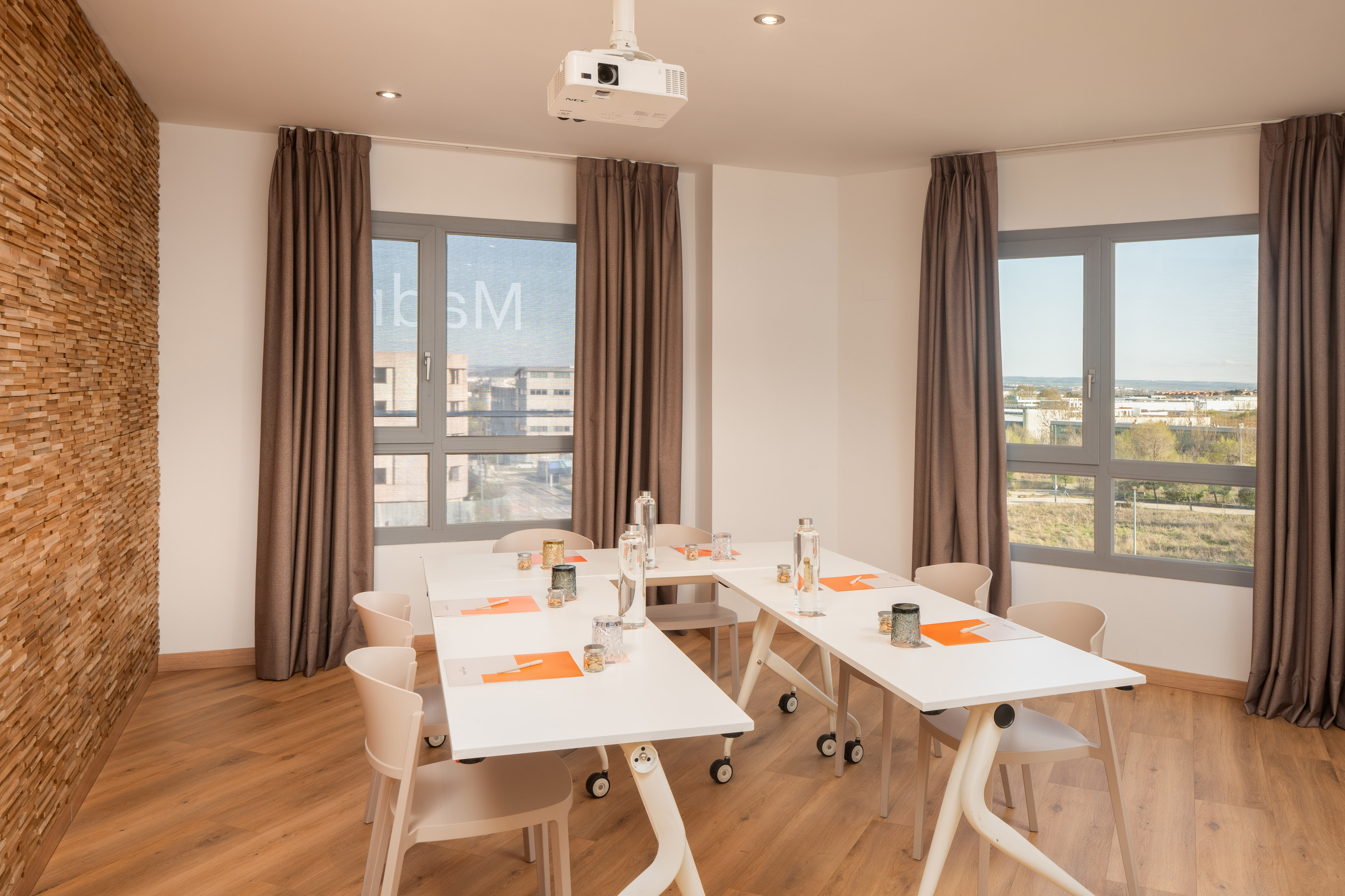 Modern meeting room with projector, white tables, large windows, wood wall.