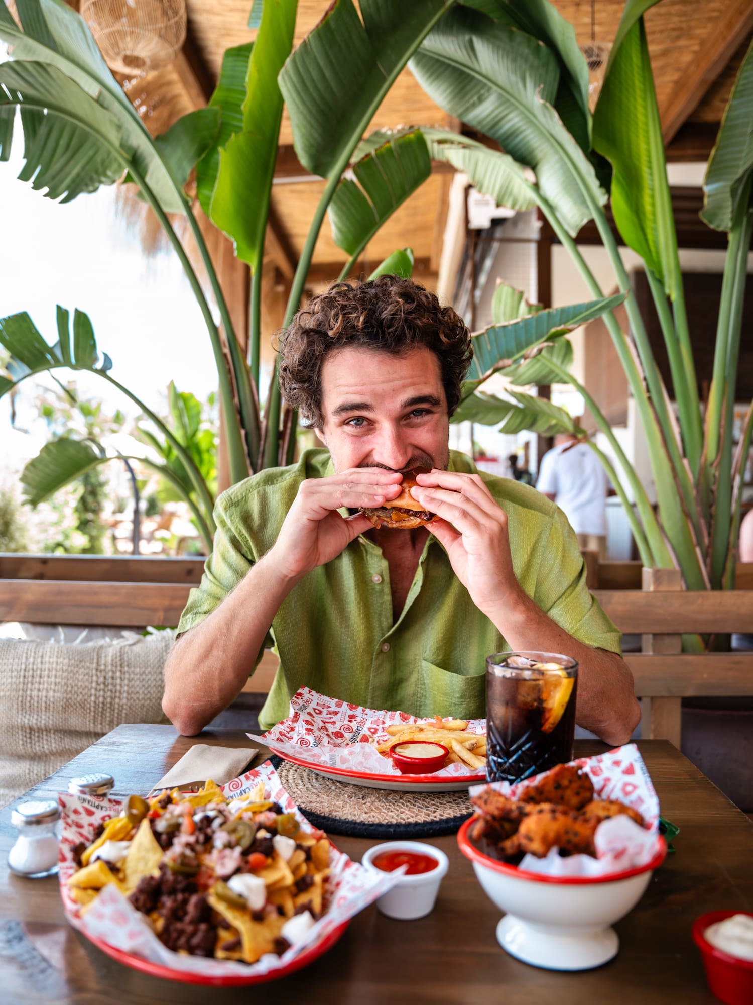Curly-haired man eating burger, fries, and nachos in tropical restaurant.