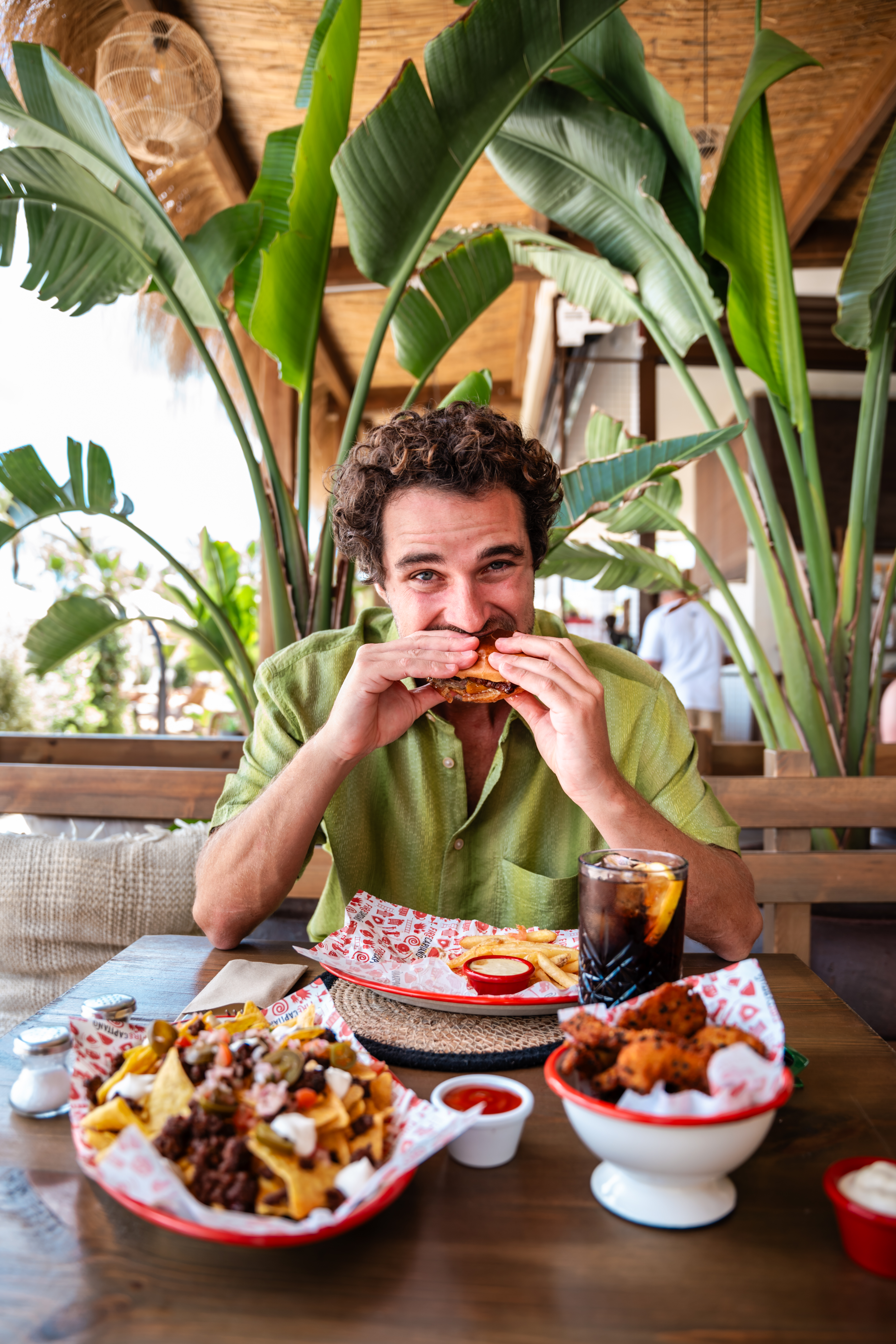 Curly-haired man eating burger, fries, and nachos in tropical restaurant.