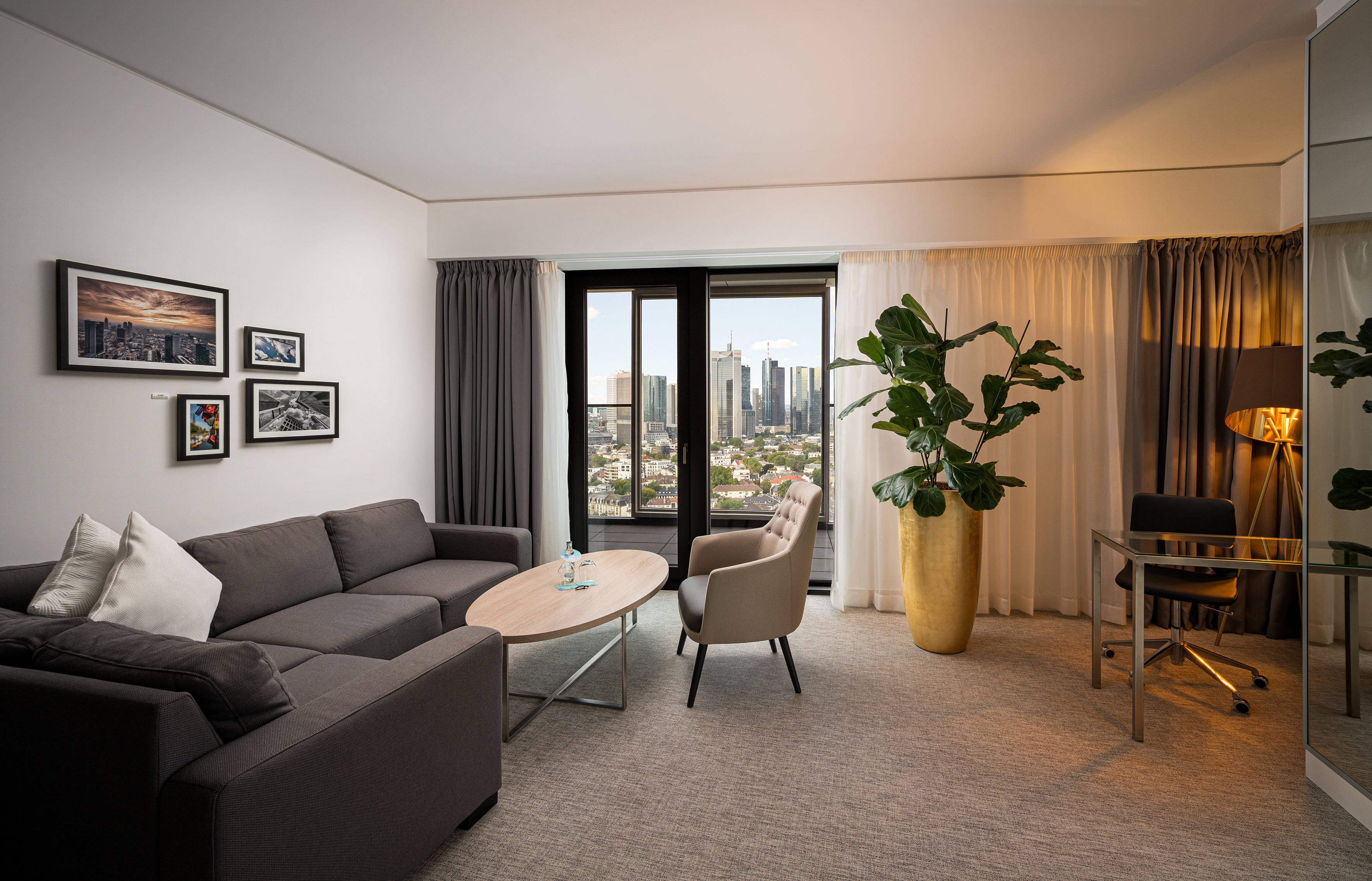 Modern hotel living room with cityscape view, grey sofa, armchair, and workspace.